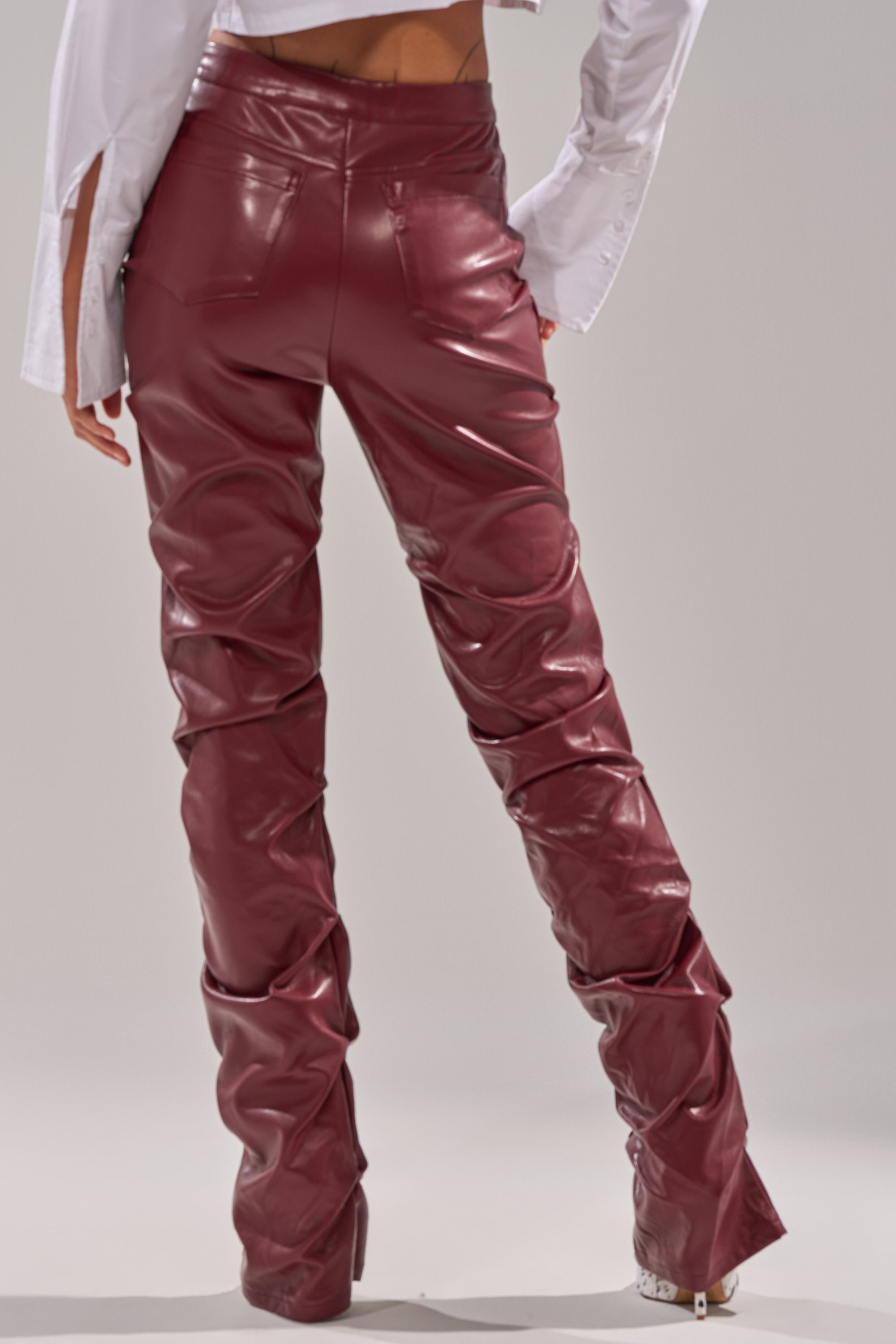 Wearing the DOWN AND DIRTY FAUX LEATHER PANT in glossy burgundy, a person stands facing away in a white shirt, highlighting the distinctive gathered texture and skinny-leg style at the back. 4