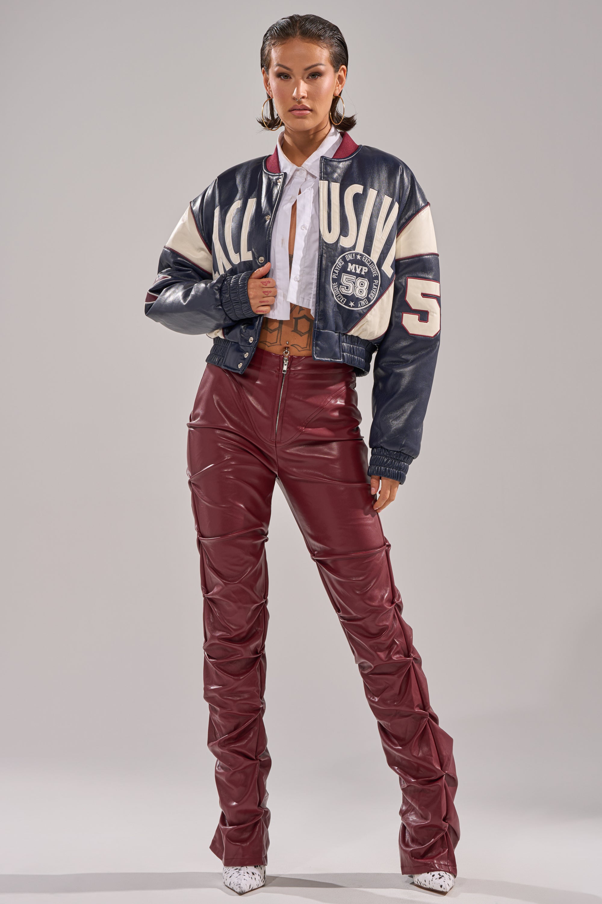 A woman stands confidently against a gray background, wearing a shiny varsity jacket, white button-up shirt, white heeled boots, and the DOWN AND DIRTY FAUX LEATHER PANT—high-waisted burgundy faux leather with a skinny-leg silhouette. 6
