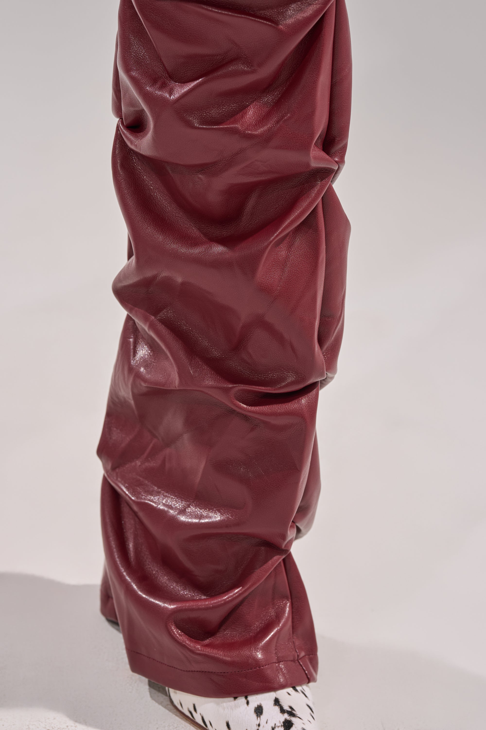 A close-up of a leg in shiny, wrinkled burgundy DOWN AND DIRTY FAUX LEATHER PANT with a skinny-leg silhouette, paired with a white and black spotted shoe against a neutral background. 7
