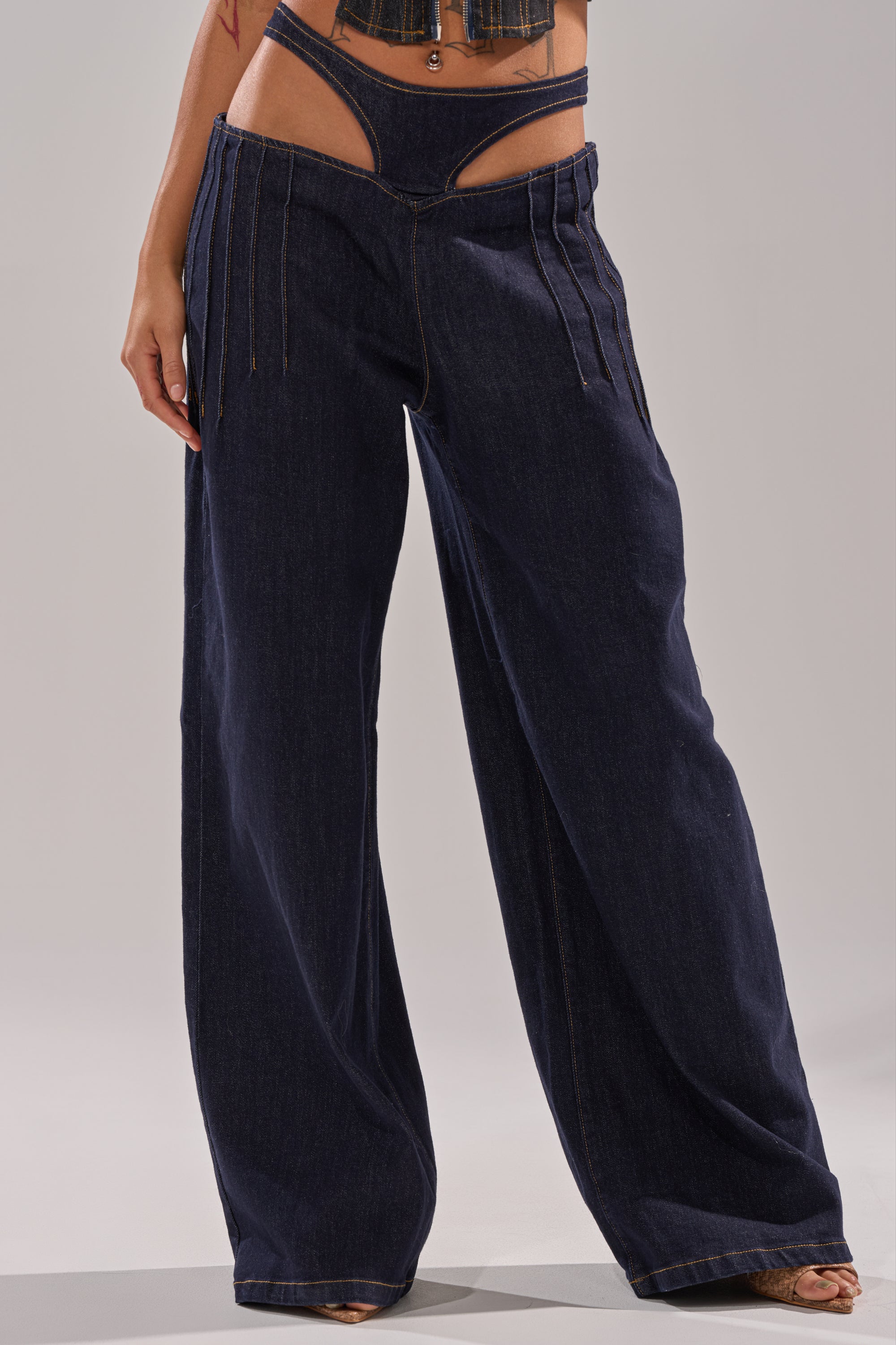 Someone wears SEXY CAN I RELAXED DENIM, featuring dark blue wide-leg pants with a low-rise cutout waistband; paired with open-toe heels, only the lower torso and legs are shown. 1