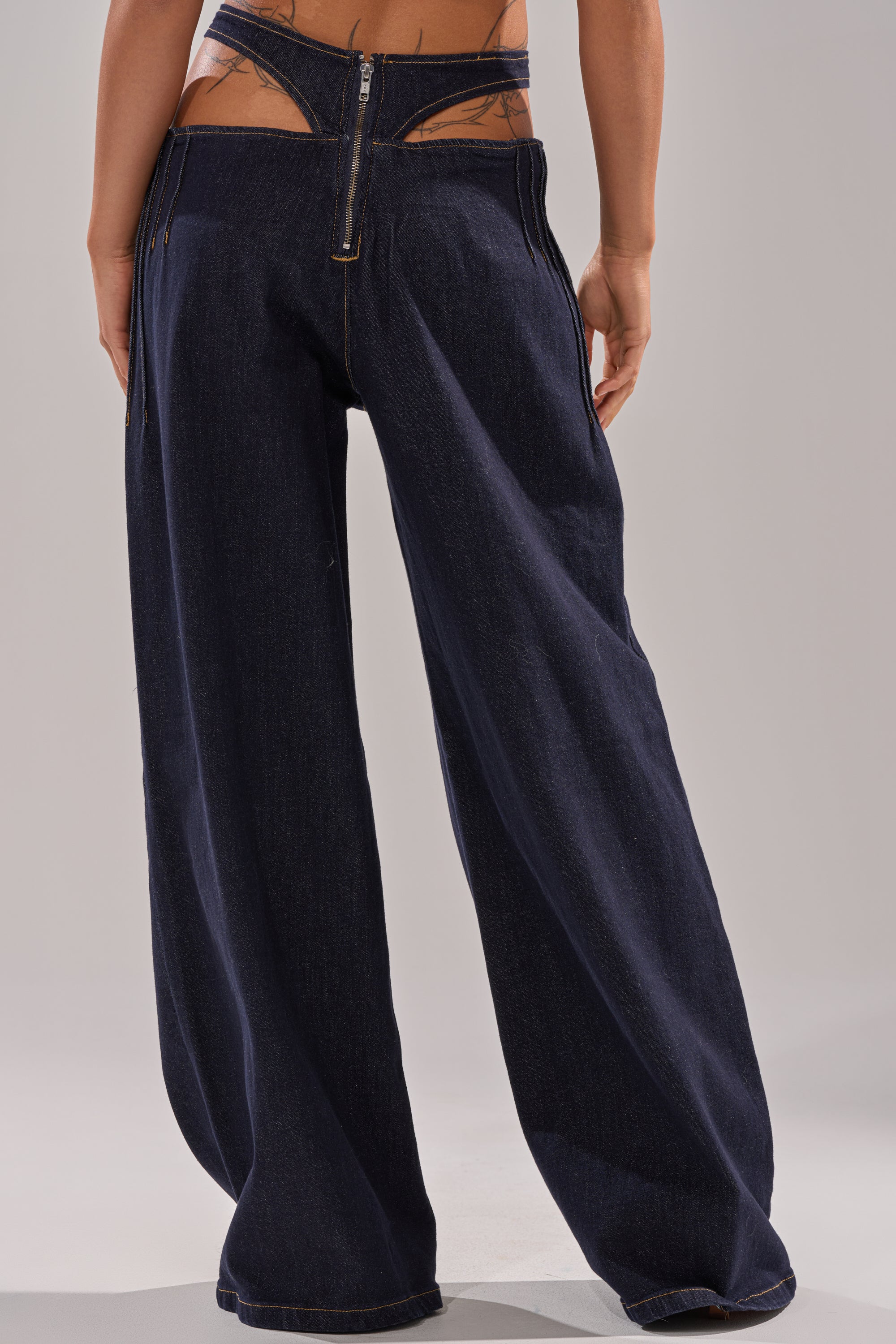 A person faces away, wearing the SEXY CAN I RELAXED DENIM—Y2K-inspired high-waisted, wide-leg dark jeans with a cut-out waistband revealing part of their lower back and hip. 3