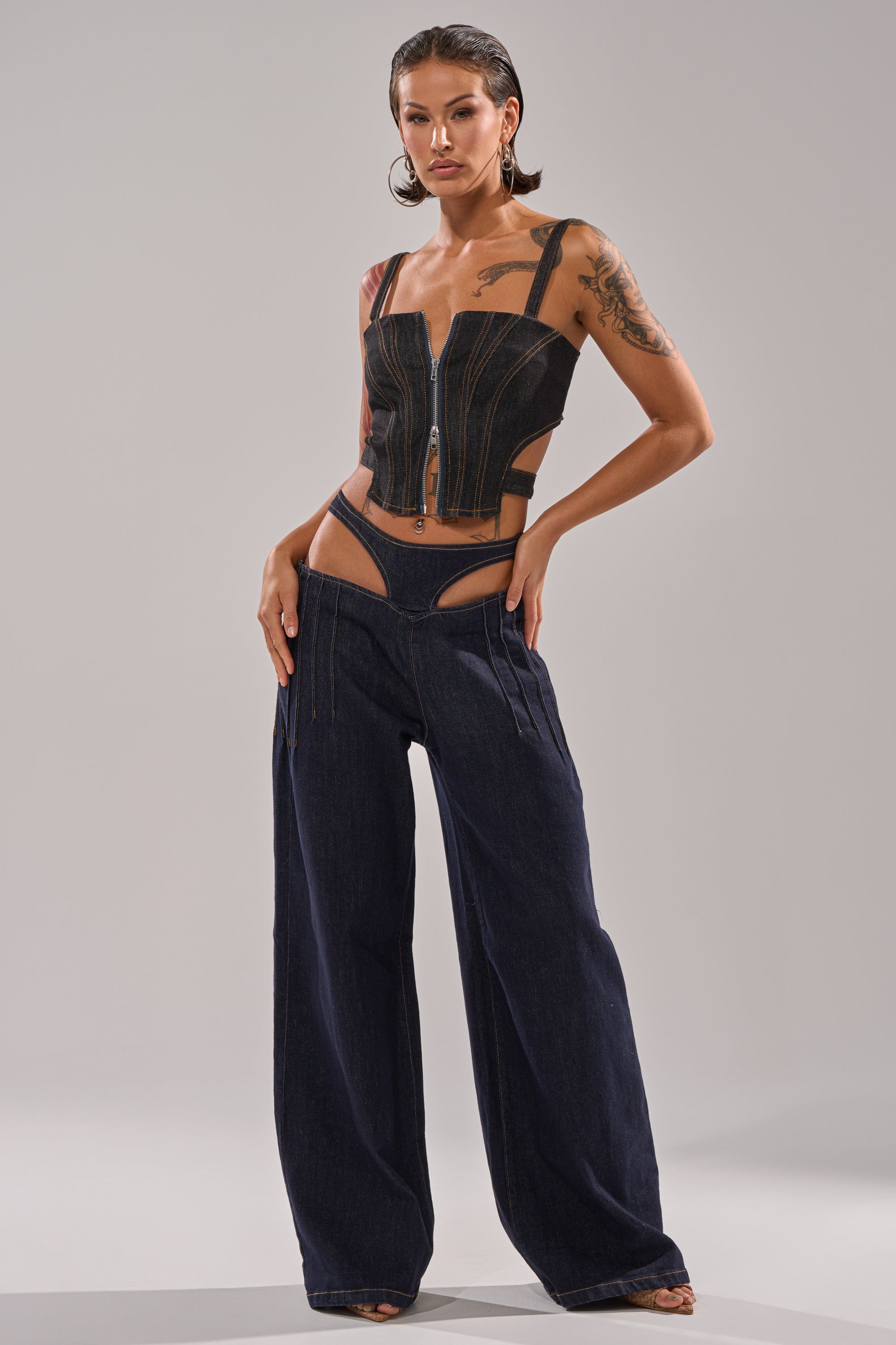 A woman poses confidently in the SEXY CAN I RELAXED DENIM, a Y2K-inspired corset-style zip-up top paired with low-rise wide-leg jeans featuring a bold cut-out waistband. She has wet-look hair and visible arm tattoos. 4