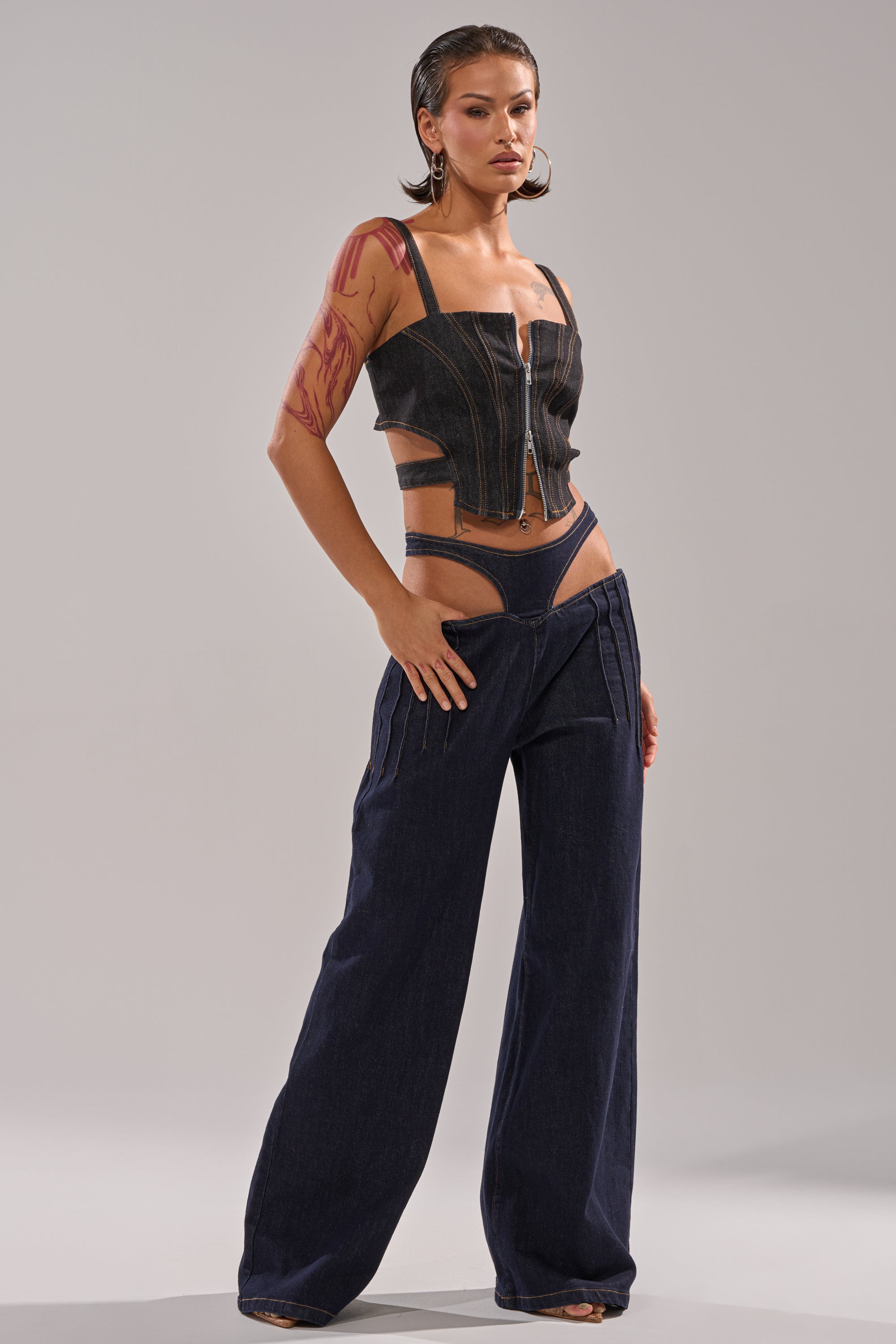 A woman stands confidently against a plain background in the SEXY CAN I RELAXED DENIM set: a sleeveless, fitted black denim corset top with front zipper, paired with matching low-rise wide-leg jeans featuring hip cutouts. 5