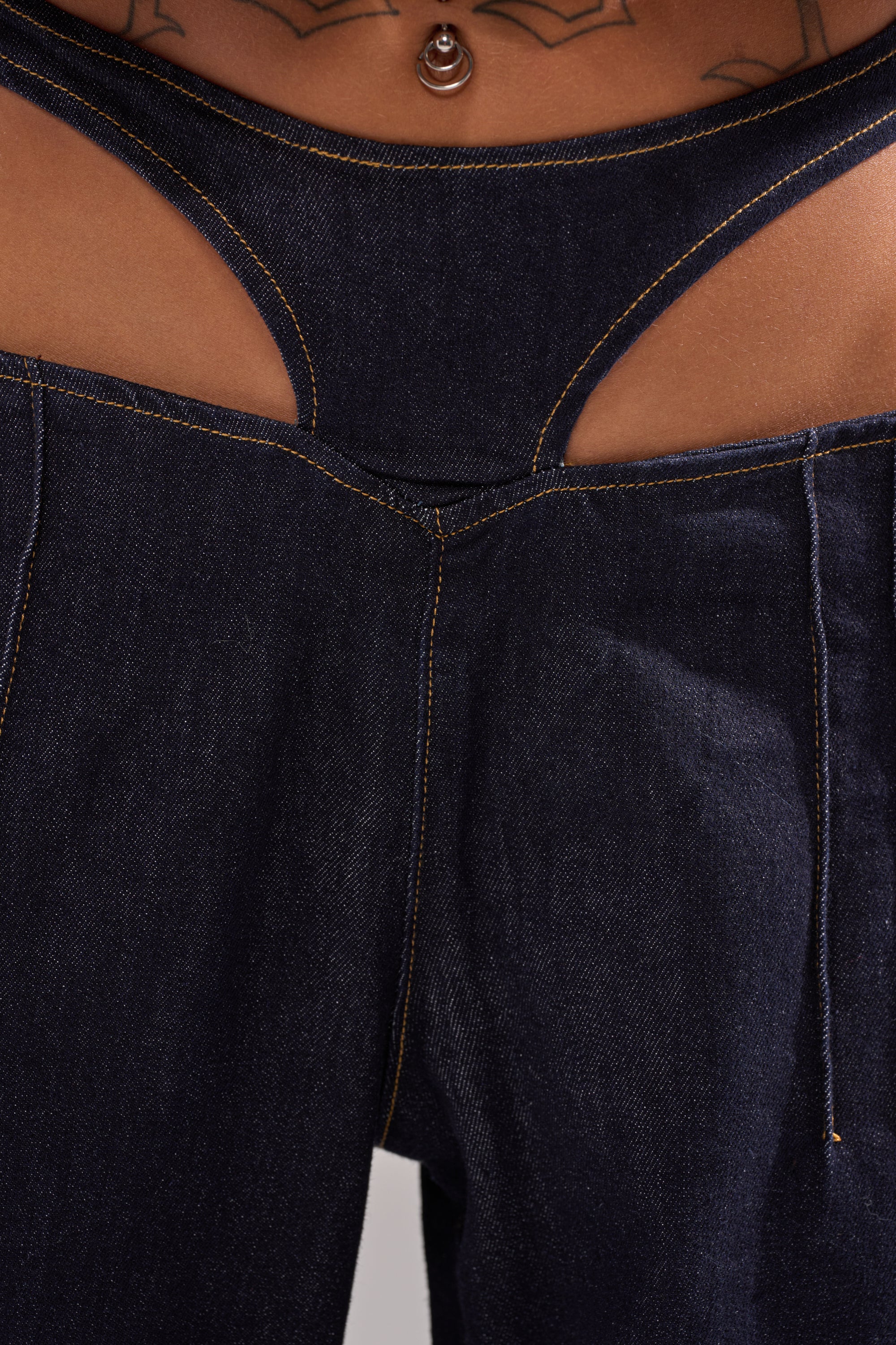 Close-up of someone wearing SEXY CAN I RELAXED DENIM, featuring a cutout waistband above low-rise wide-leg jeans. Tattoos and a navel piercing complement the edgy style. 7