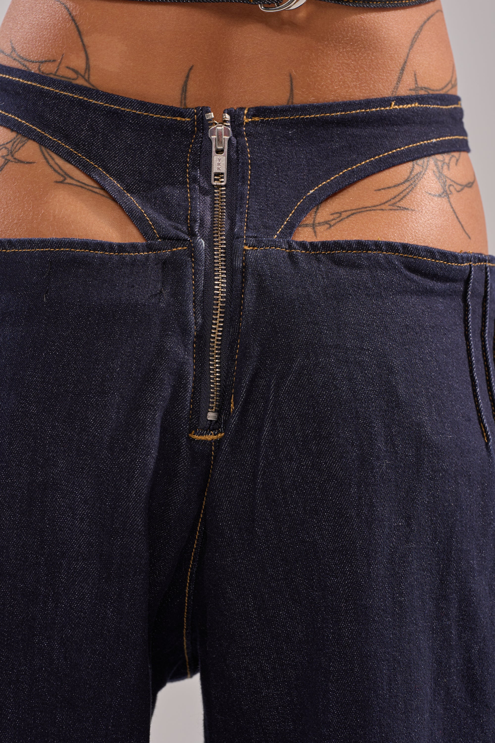 Close-up of a person wearing SEXY CAN I RELAXED DENIM: Y2K-inspired dark blue jeans with a cut-out waistband, visible zipper, decorative stitching, and wide-leg silhouette. Tattooed skin shows above the relaxed fit. 8