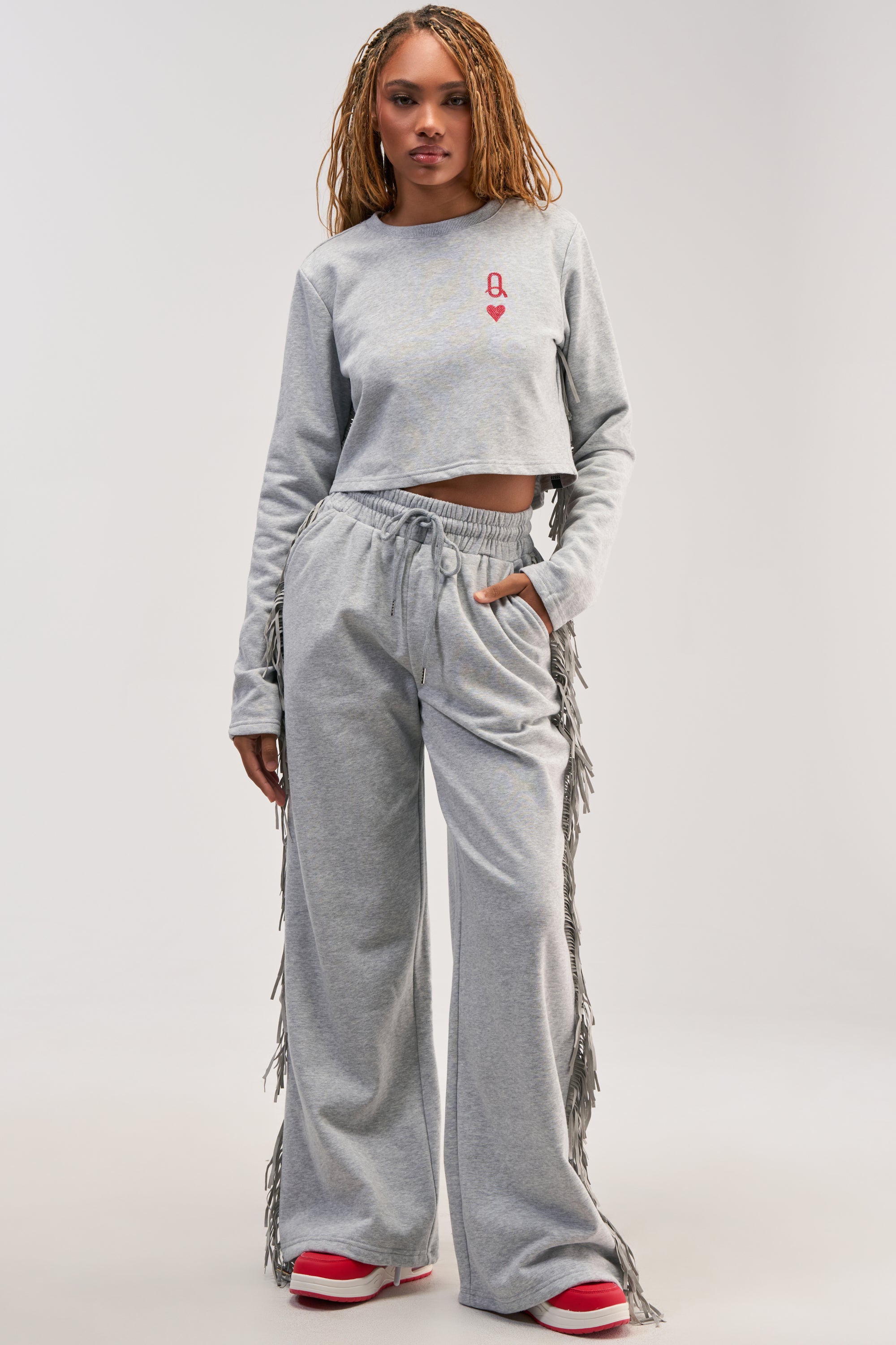 A woman stands facing forward against a plain white background, wearing the PLAYING CARDS WIDE LEG JOGGER IN HEATHER GREY paired with a gray crop top. She has long braided hair and completes her relaxed yet stylish look with red and white sneakers.