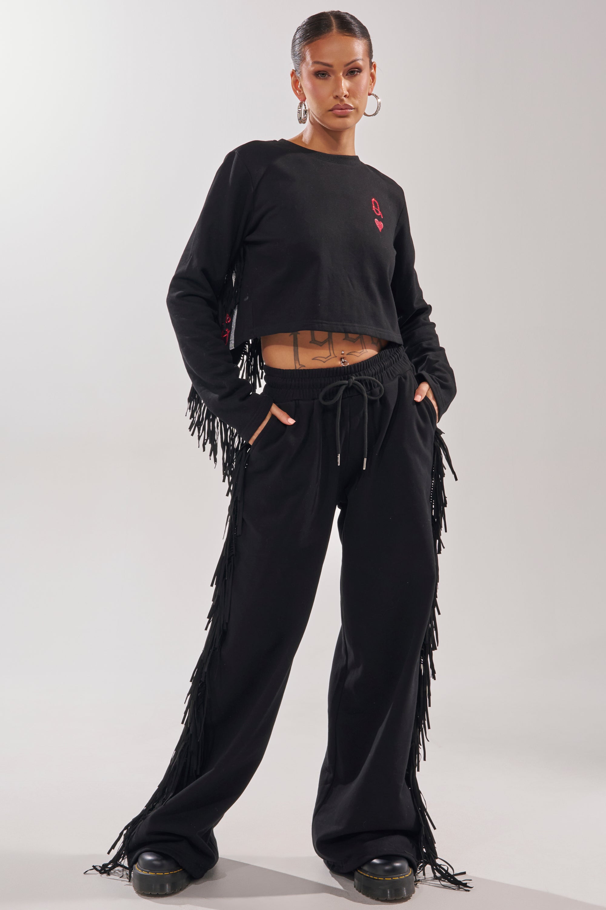 A woman stands confidently in the PLAYING CARDS WIDE LEG JOGGER and a matching long-sleeve crop top, her hands in her pockets. Hoop earrings and black shoes complete her look, highlighting the stylish fringe detailing down the jogger’s sides. 0