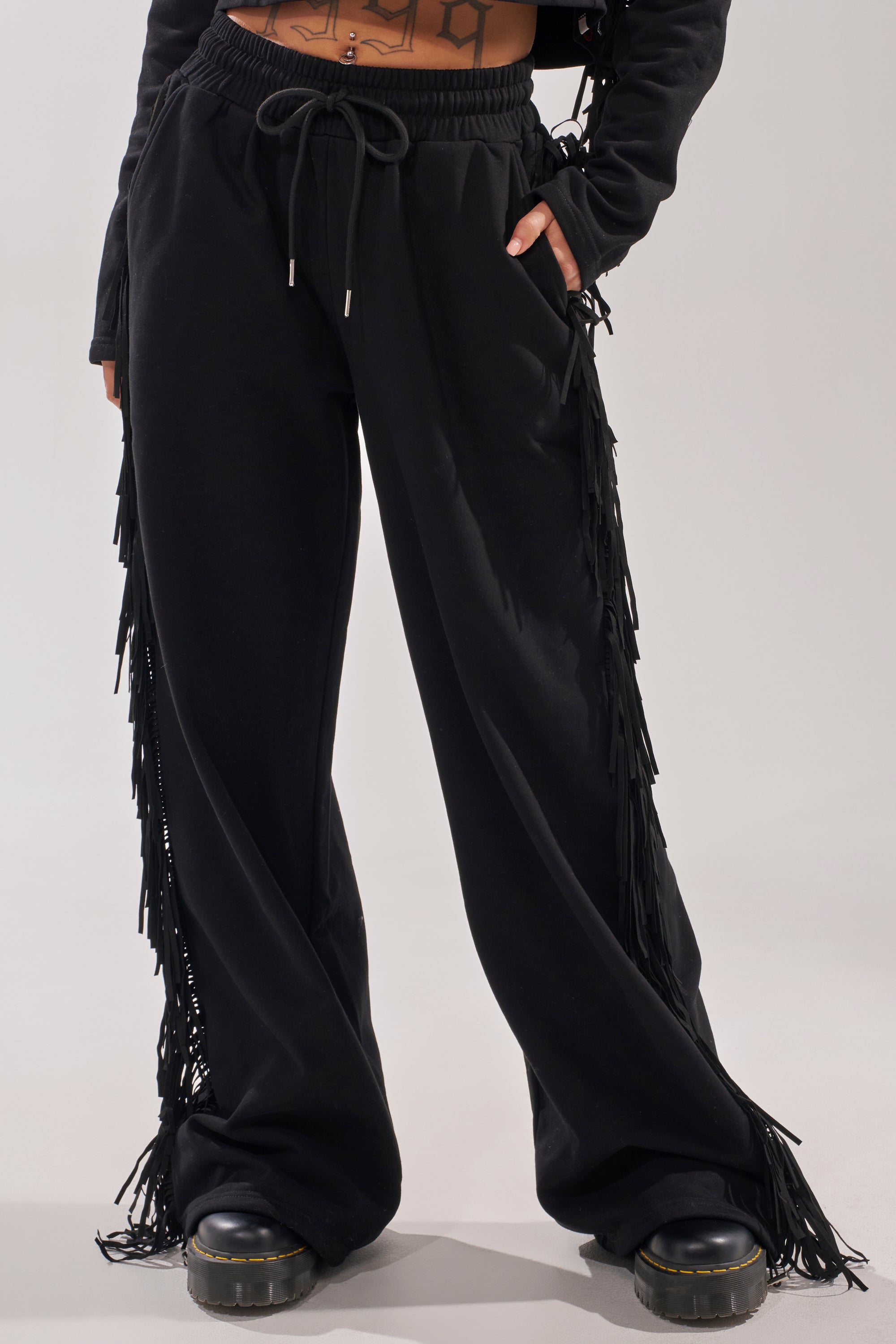 A person wearing PLAYING CARDS WIDE LEG JOGGER—black, wide-leg cotton-blend joggers with fringe detailing and a drawstring waist—paired with platform shoes, hands in pockets. Only the lower torso and legs are visible. 1