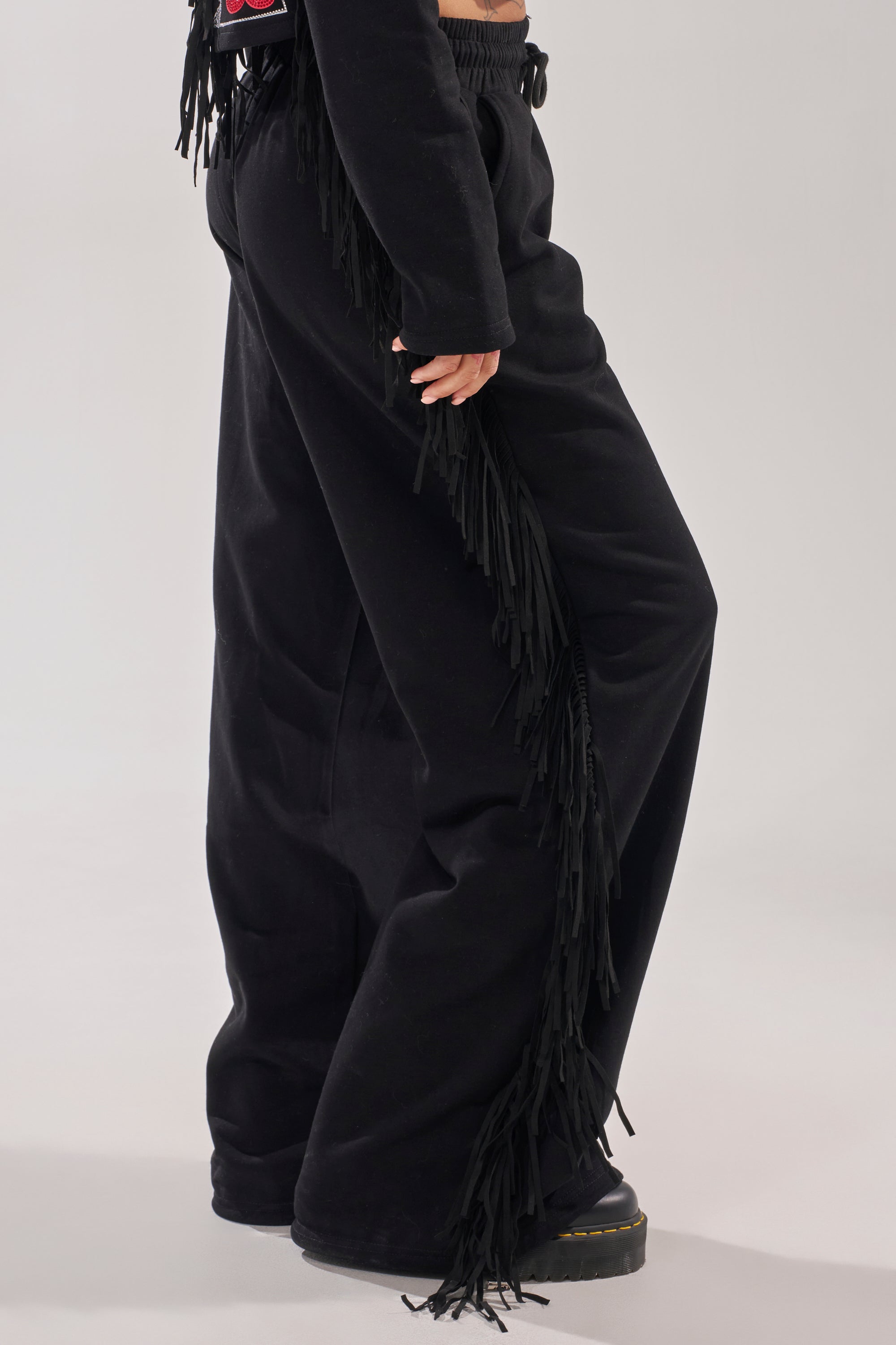 A person wearing PLAYING CARDS WIDE LEG JOGGER with fringe on the side seams, paired with black shoes and a matching black top featuring fringe, stands against a plain light background. 3