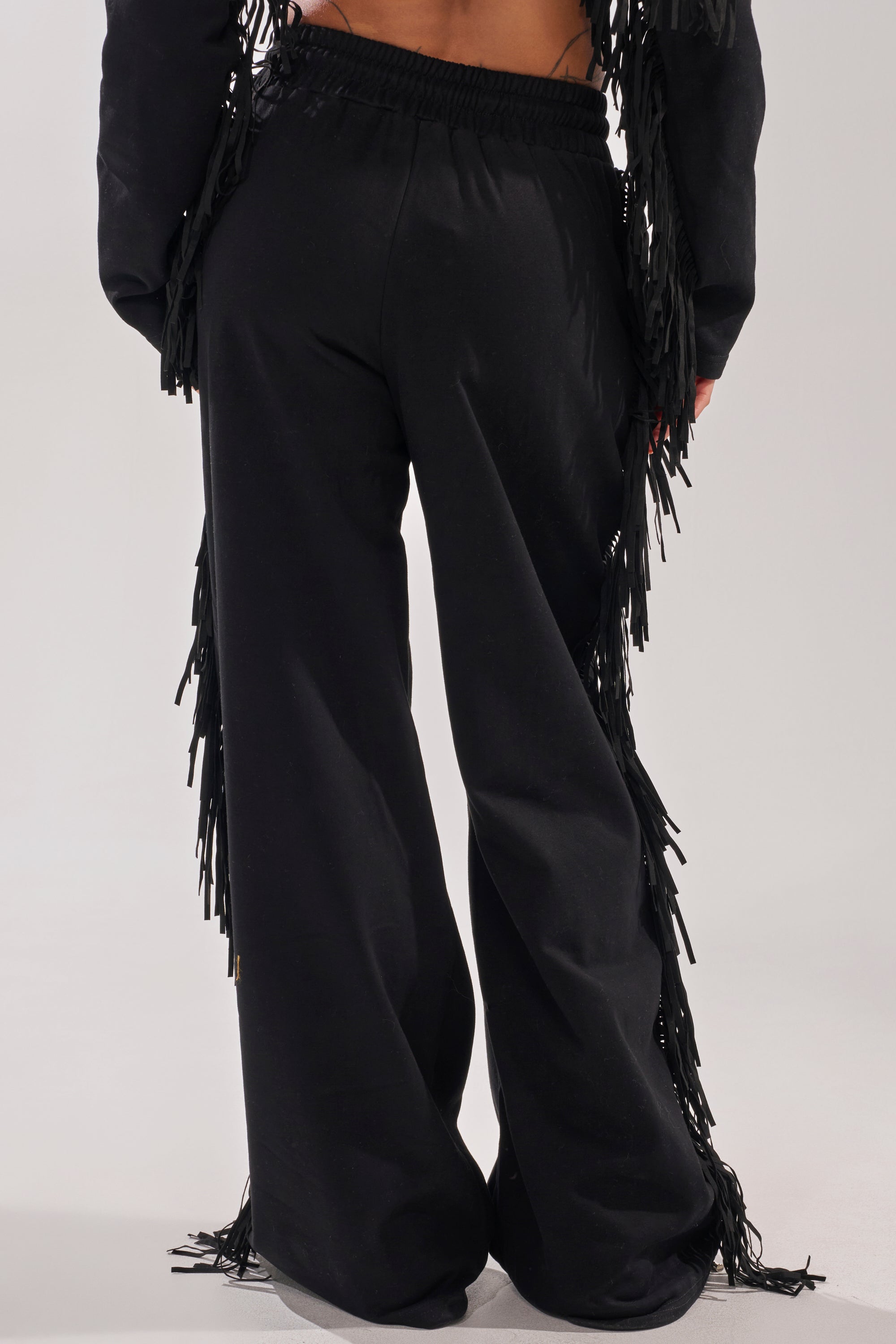 A person stands facing away, wearing the PLAYING CARDS WIDE LEG JOGGER—black joggers with long fringe along the outer seams, featuring a loose fit and an elastic waistband for comfort. 4