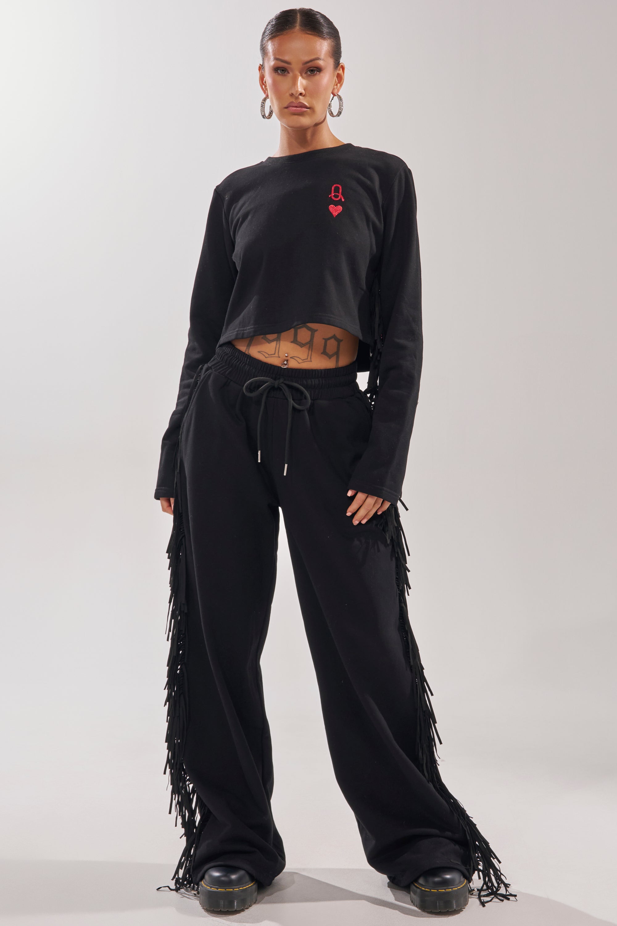 A person stands confidently in the PLAYING CARDS WIDE LEG JOGGER, paired with a black long-sleeve crop top with a red logo, hoop earrings, and black shoes. Their hair is pulled back, midriff shows geometric tattoos. Background is plain. 5