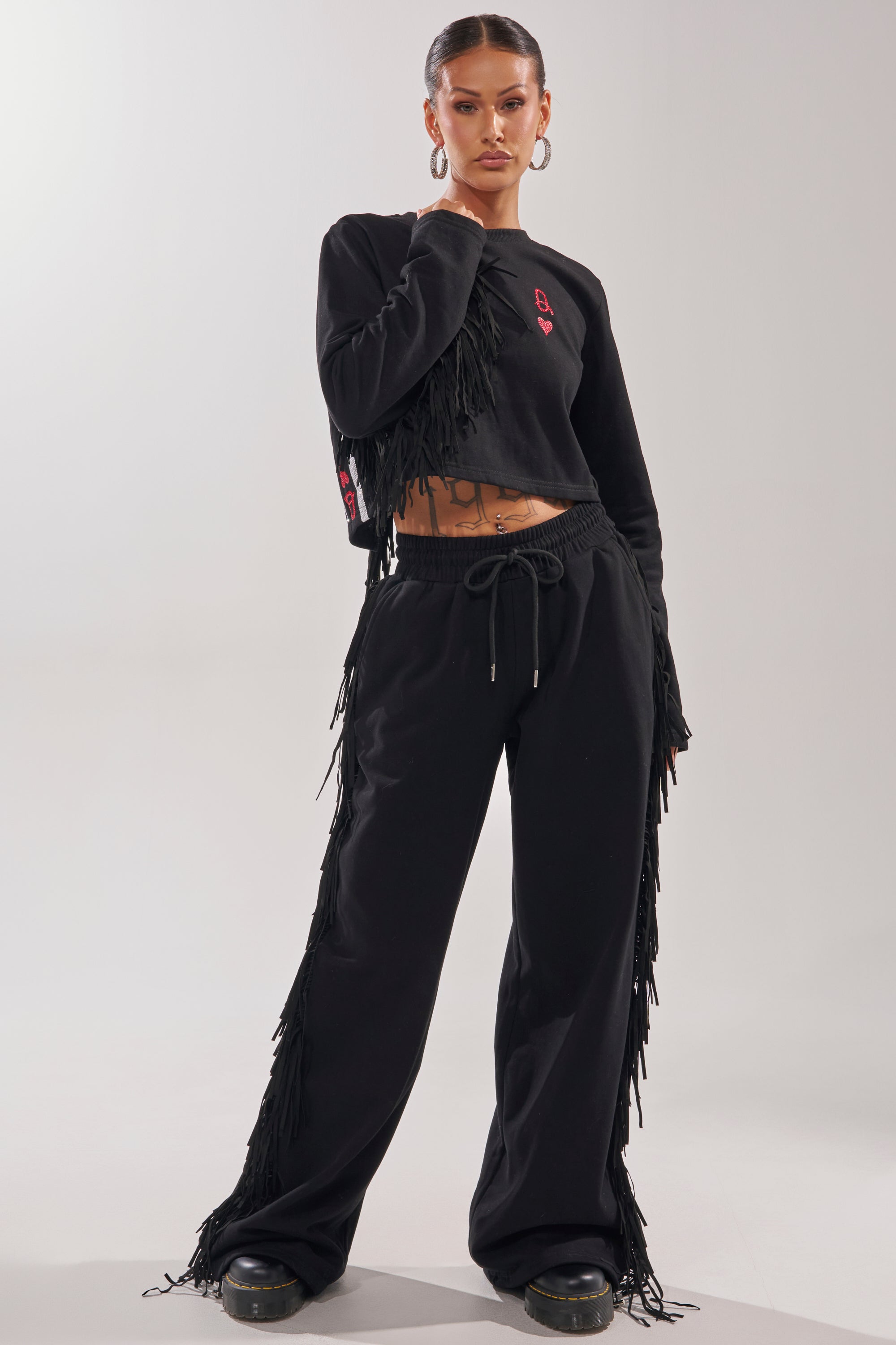 A woman poses confidently in the PLAYING CARDS WIDE LEG JOGGER with a matching fringe-trimmed crop top, hoop earrings, slicked-back hair, and black shoes against a simple light background. 6