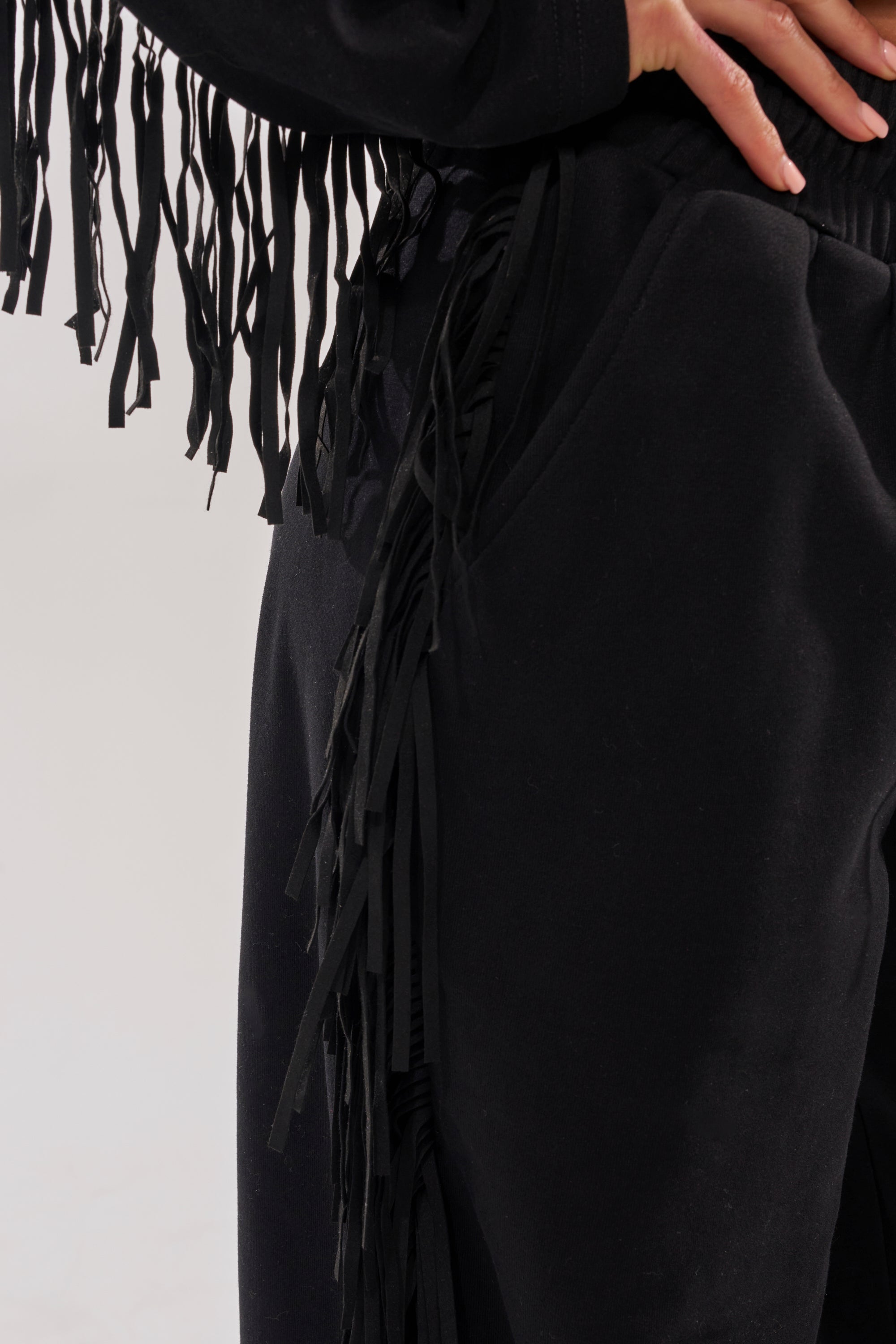 Close-up of a person wearing the PLAYING CARDS WIDE LEG JOGGER and matching top, both featuring long, thin fringe details. One hand is on the hip, drawing attention to the lower torso and upper legs. 7