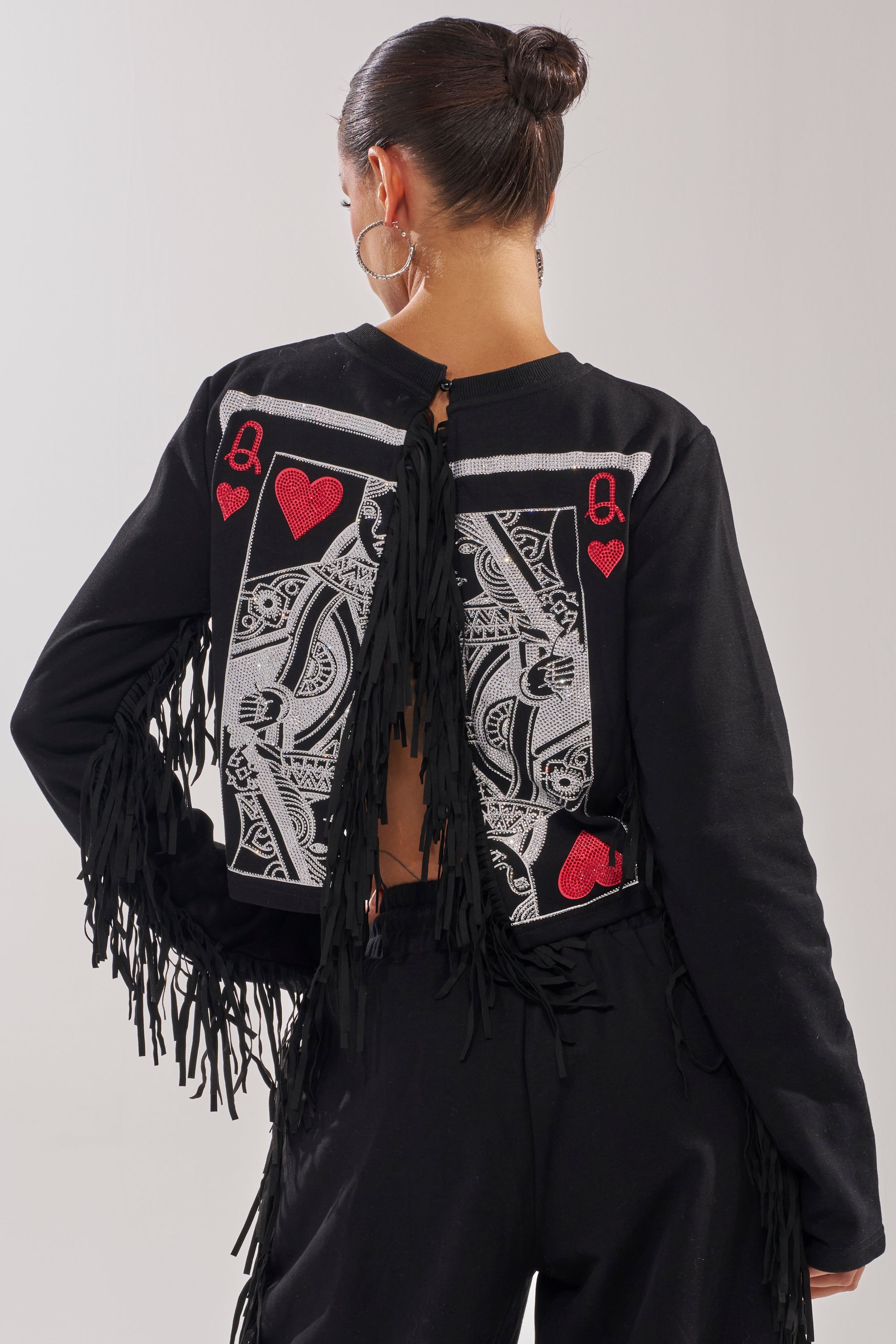 A woman with her hair in a bun wears the PLAYING CARDS CROP SWEATSHIRT, featuring a bold playing card design on the back, black fringe on the sleeves, and an open back for a modern twist. 0