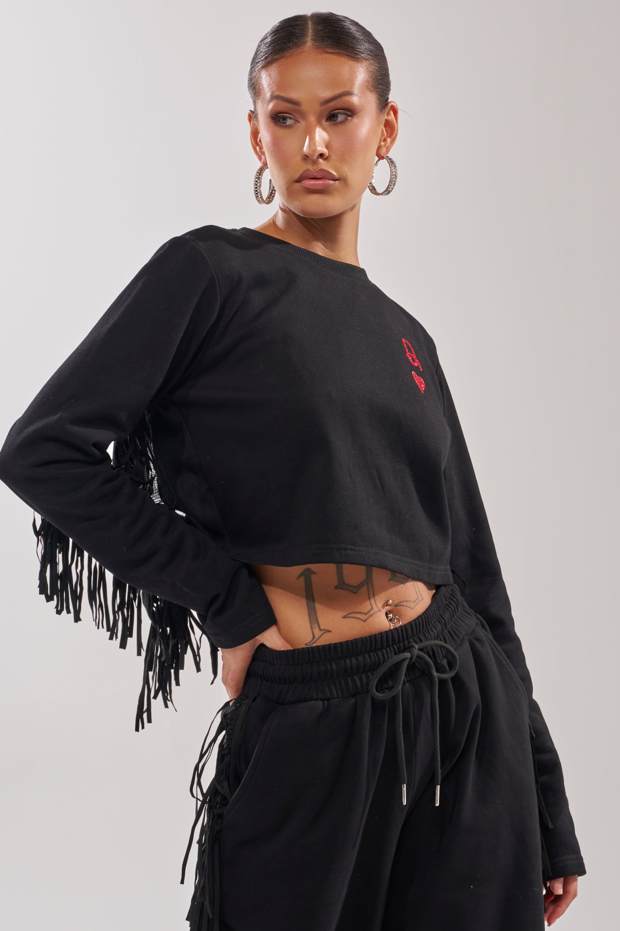 Wearing hoop earrings, a woman poses with one hand on her hip in the PLAYING CARDS CROP SWEATSHIRT and matching black drawstring pants, showing off her abdomen tattoo. 1