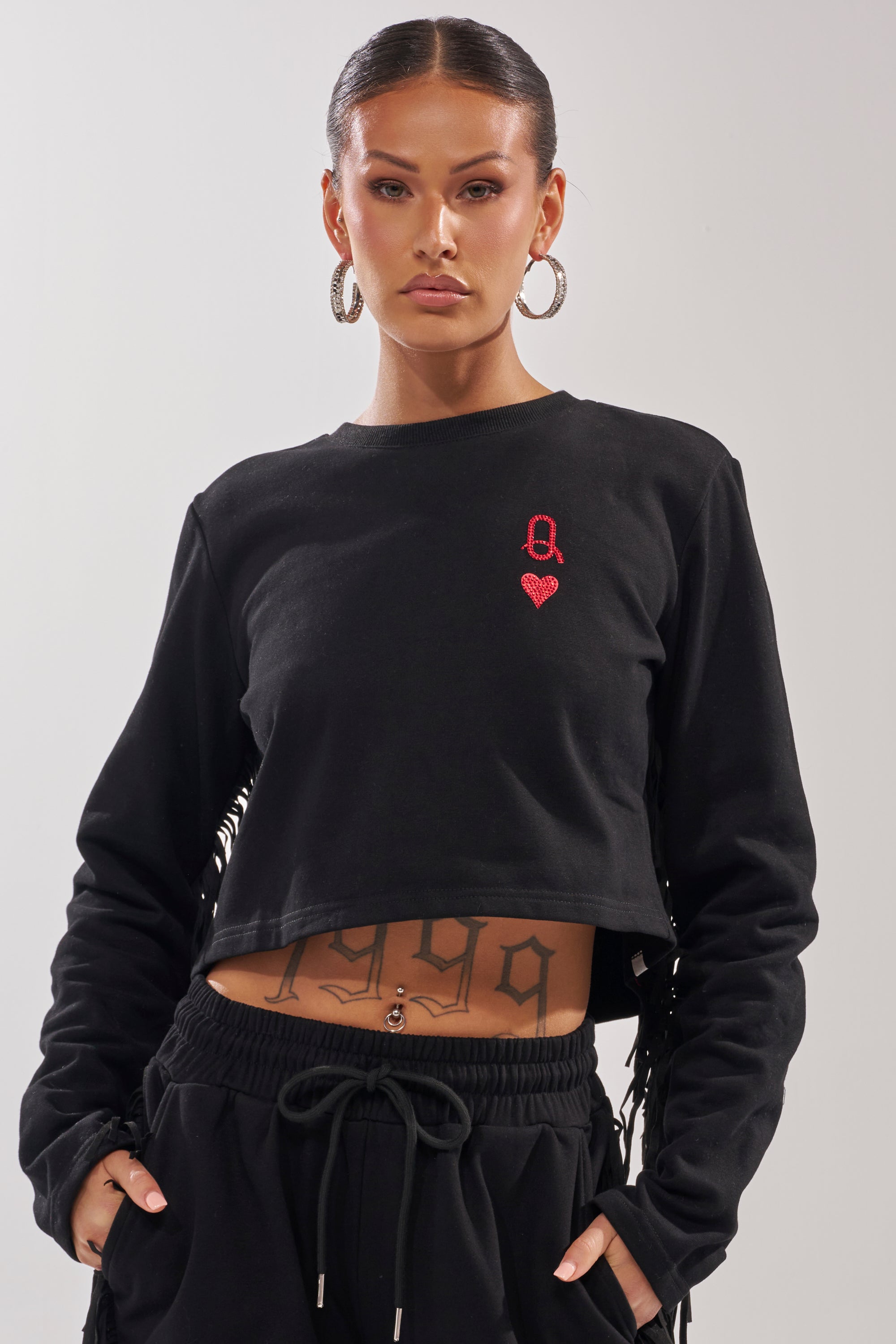 A person with hoop earrings and a nose ring wears the PLAYING CARDS CROP SWEATSHIRT—black, featuring an embroidered red "A" and heart—paired with black drawstring pants, and has "1999" tattooed on their lower abdomen. 2