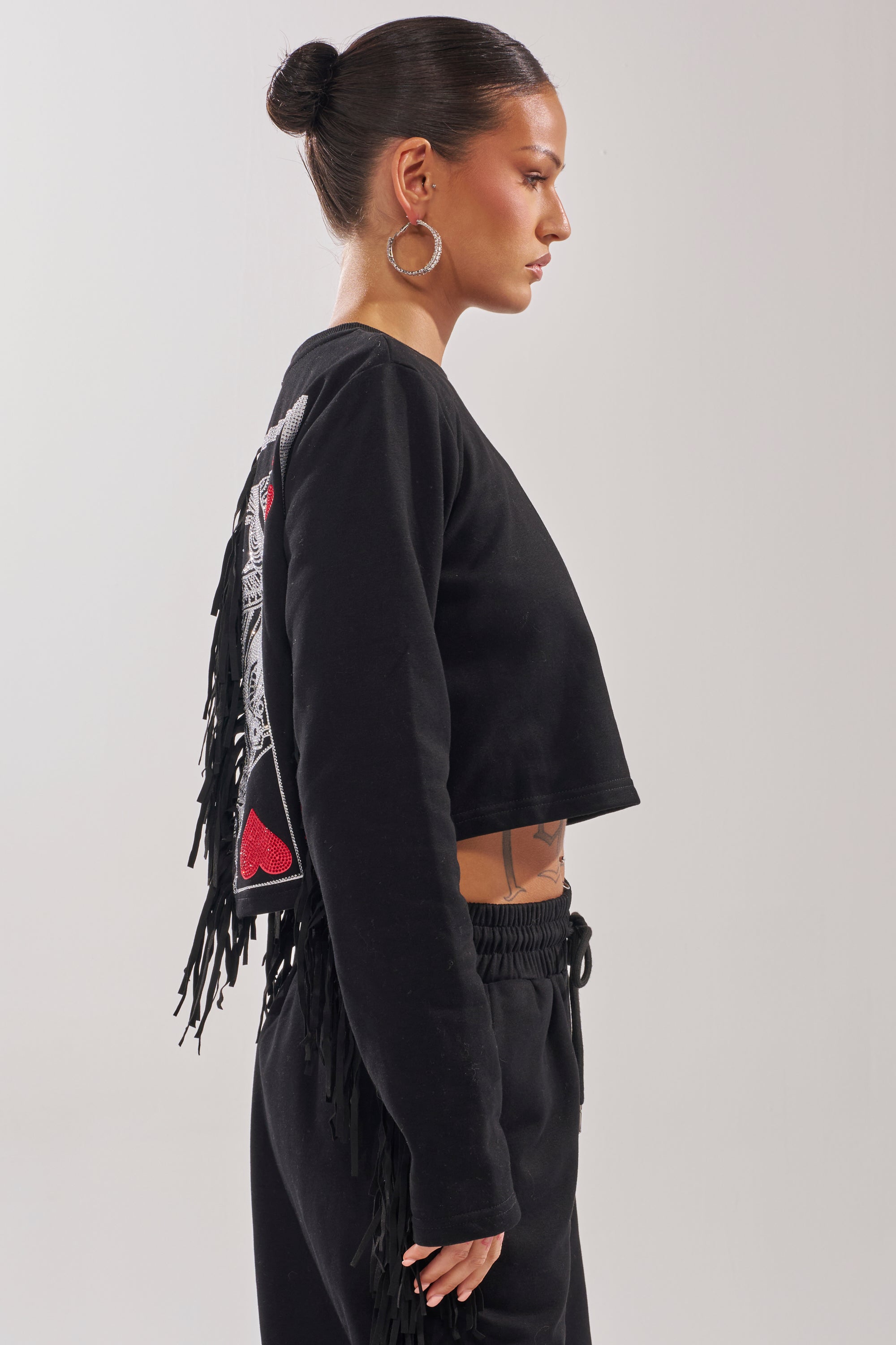 A woman with her hair in a bun wears the PLAYING CARDS CROP SWEATSHIRT— a black, long-sleeve cropped sweater with fringe trim and playful back patches—paired with matching black pants, standing in profile against a plain background. 3