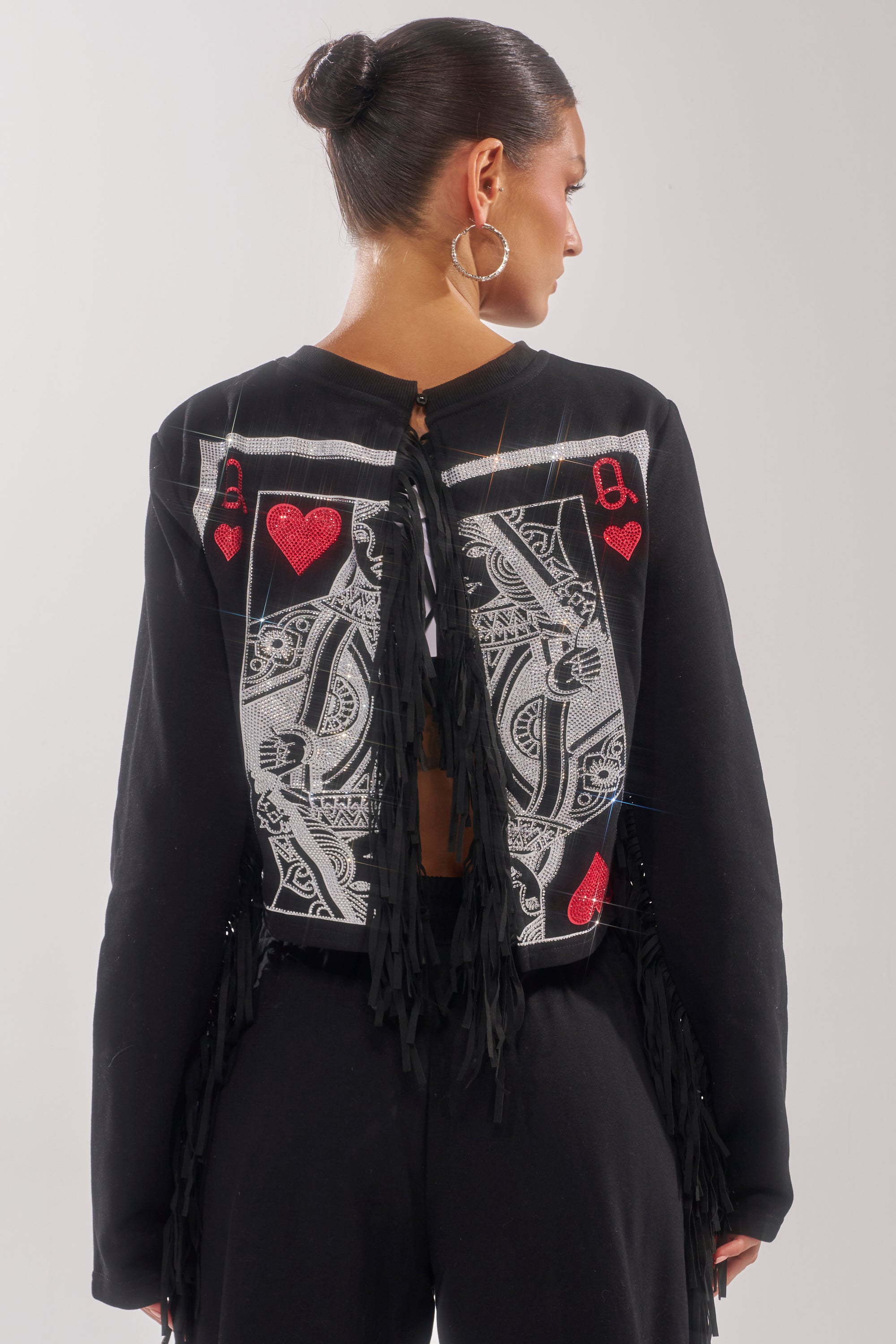 A woman with dark hair in a bun and large hoop earrings, wearing the PLAYING CARDS CROP SWEATSHIRT with red accents and fringe trim on the back, stands facing away from the camera. 4