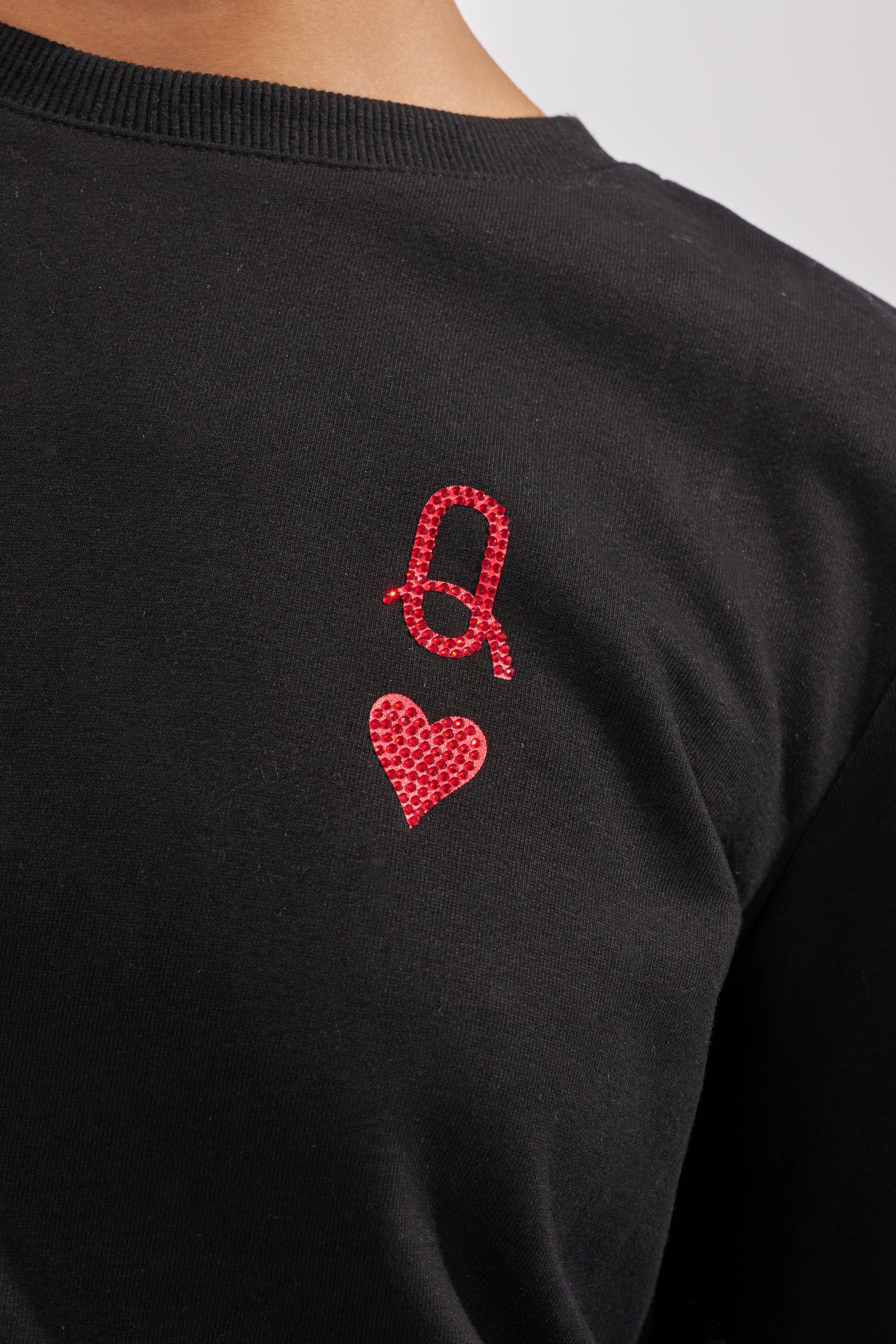 A close-up of the back of the PLAYING CARDS CROP SWEATSHIRT, featuring red rhinestones forming a loop with a tail above a heart, echoing a Queen of Hearts motif. 8