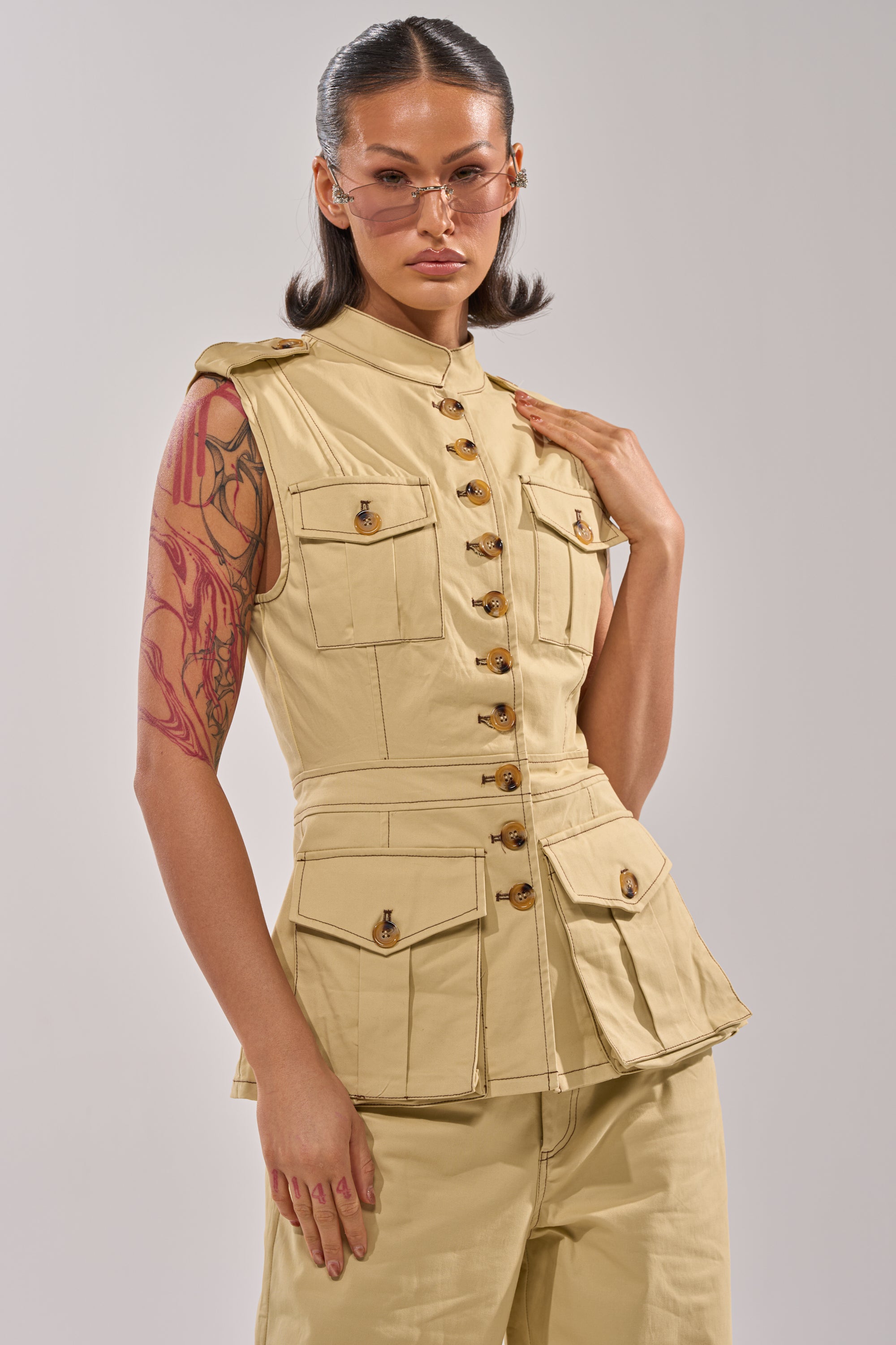 A model poses confidently in the DIANA VEST TOP with multiple pockets and matching pants, wearing sunglasses. Their sleek shoulder-length hair and visible arm tattoo stand out against a plain light background for a business-casual look. 0