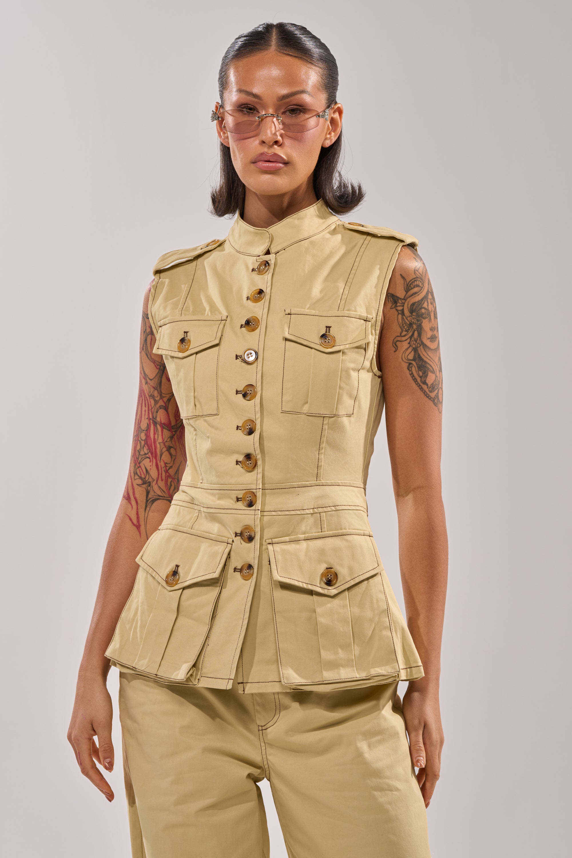 A woman with long dark hair confidently wears the DIANA VEST TOP in khaki, paired with matching beige pants and light brown sunglasses. Tattoos decorate both arms as she stands against a plain light background. 1