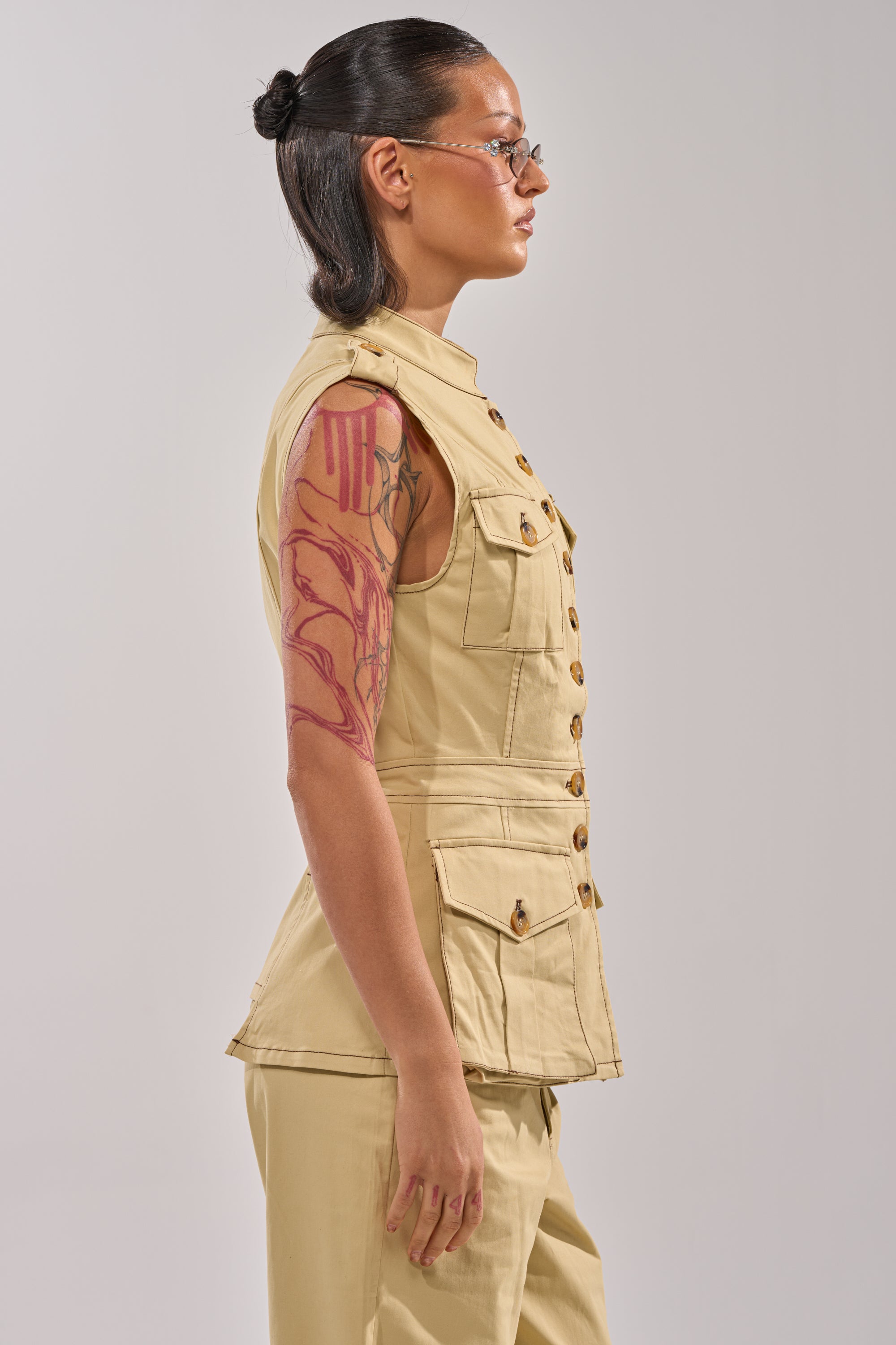 A person wearing glasses and a visible arm tattoo models the DIANA VEST TOP with matching khaki pants, standing in profile against a plain light gray background to highlight its tailored silhouette. 3