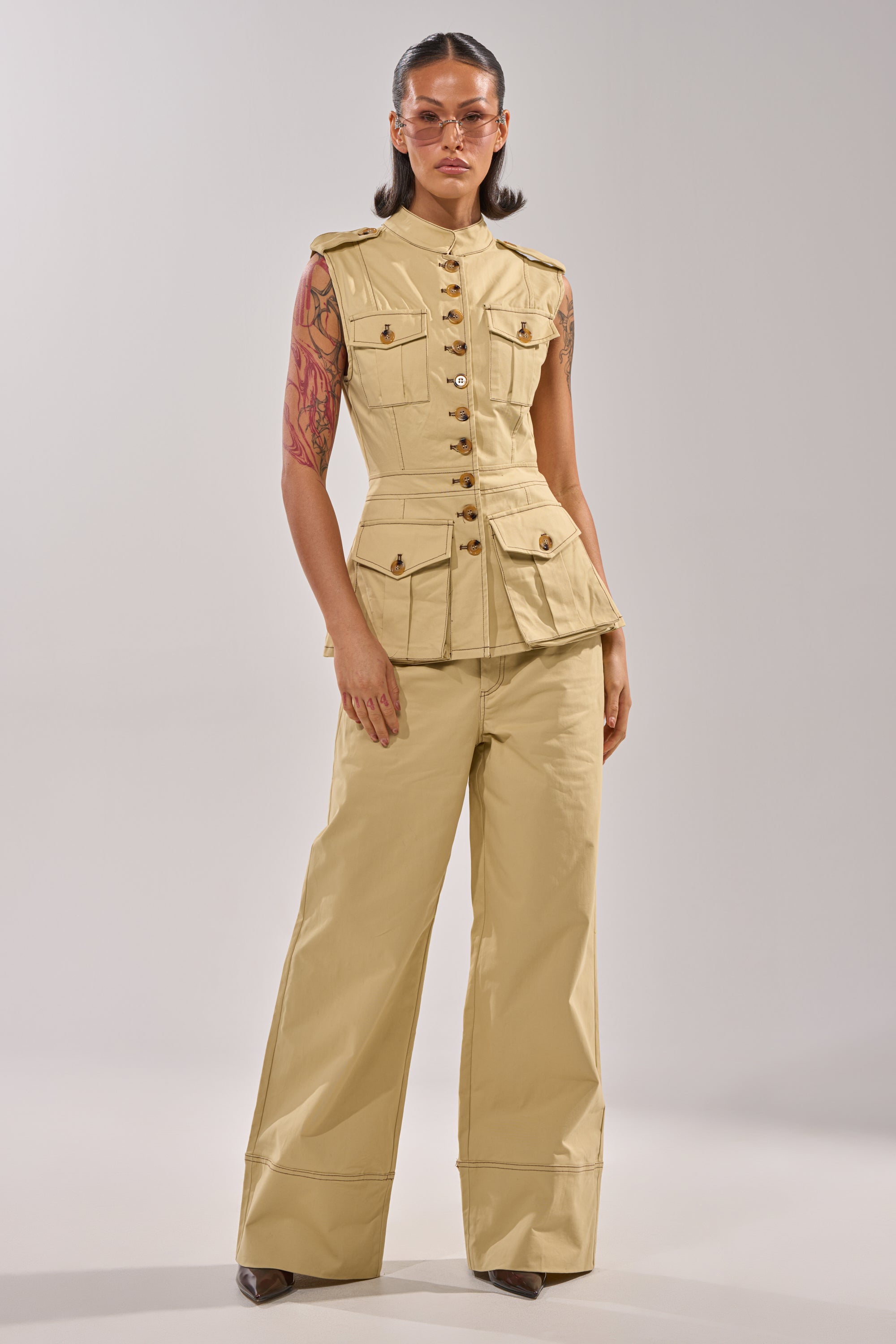 A woman in the DIANA VEST TOP and wide-leg khaki pants stands confidently against a light gray background, showcasing a tailored, business-casual look with gold buttons, sunglasses, arm tattoos, and straight dark hair. 5