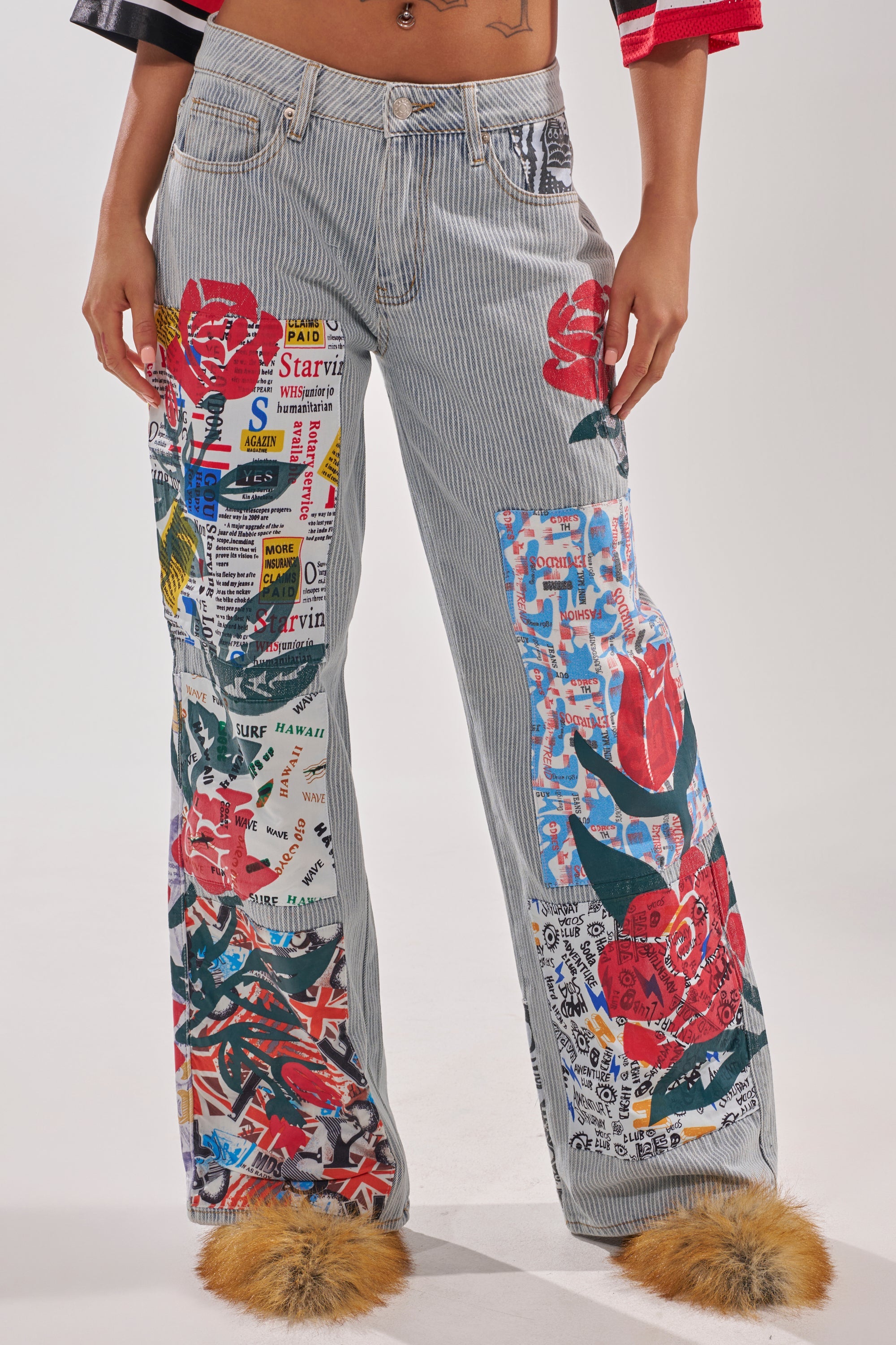 A person wears the CIAO BELLA STRIPE TROUSER, striped denim wide-leg pants adorned with colorful patches of red roses, text, and graphics, styled with fuzzy tan shoes and a partly visible cropped top. 0