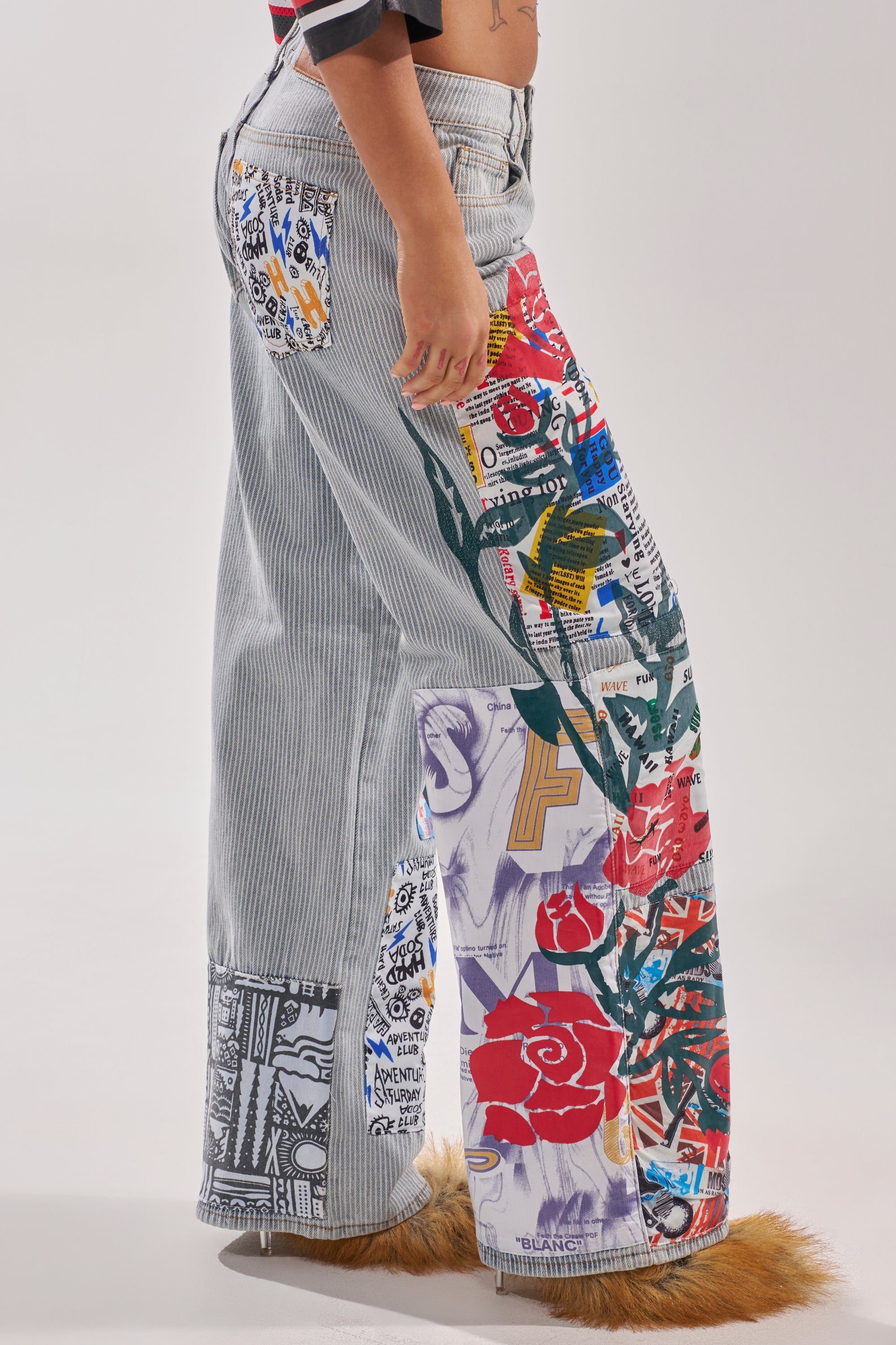 A person stands sideways against a plain background wearing the CIAO BELLA STRIPE TROUSER—high-rise, wide-leg jeans featuring vertical pinstripes, bold colorful patches, graphic prints, and furry brown ankle cuffs. 2
