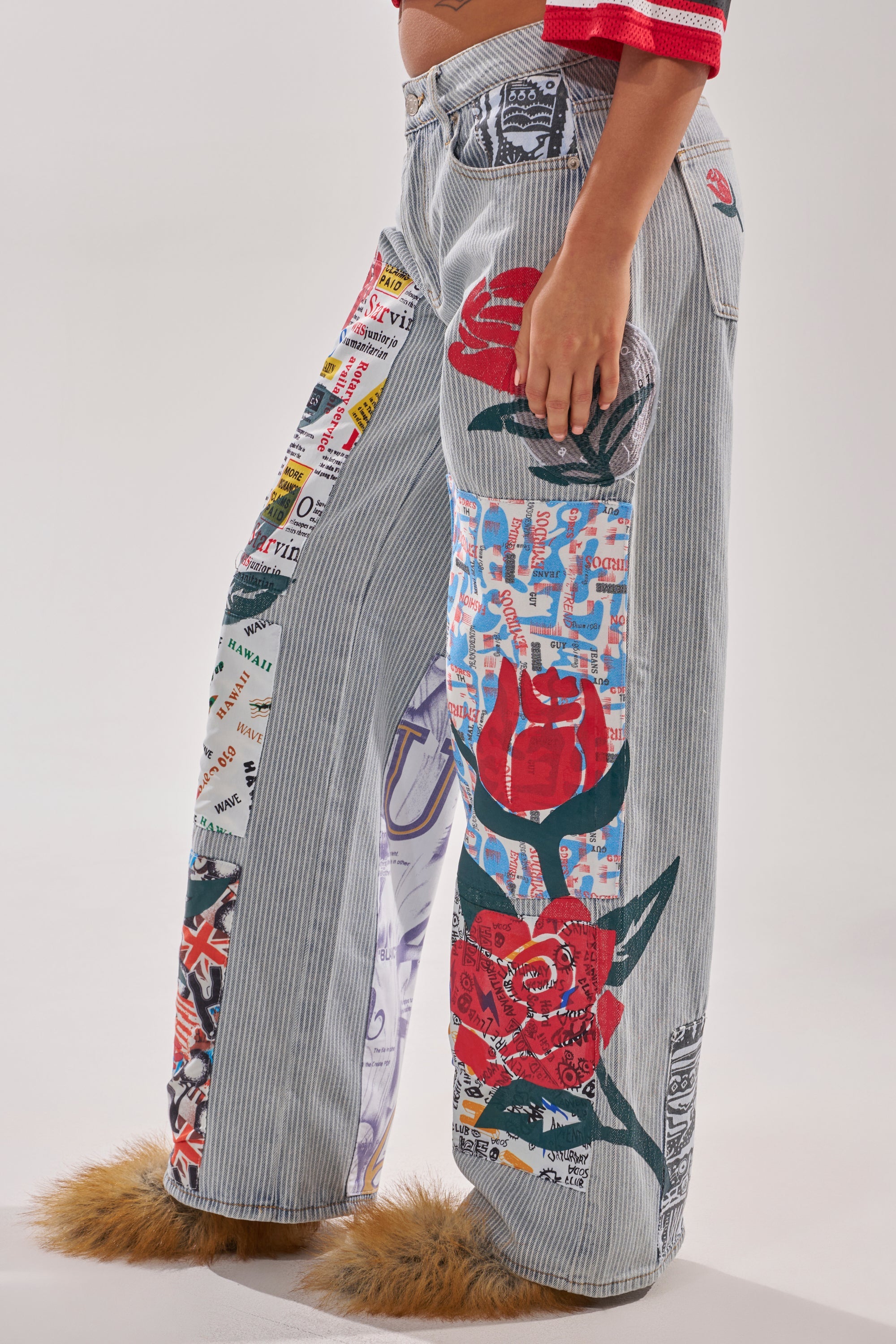 A person wearing the CIAO BELLA STRIPE TROUSER—patchwork denim with bold stripes, collage-style patches, red roses, text, and graphics—paired with tan furry slippers and a red top with white sleeve stripes. 3