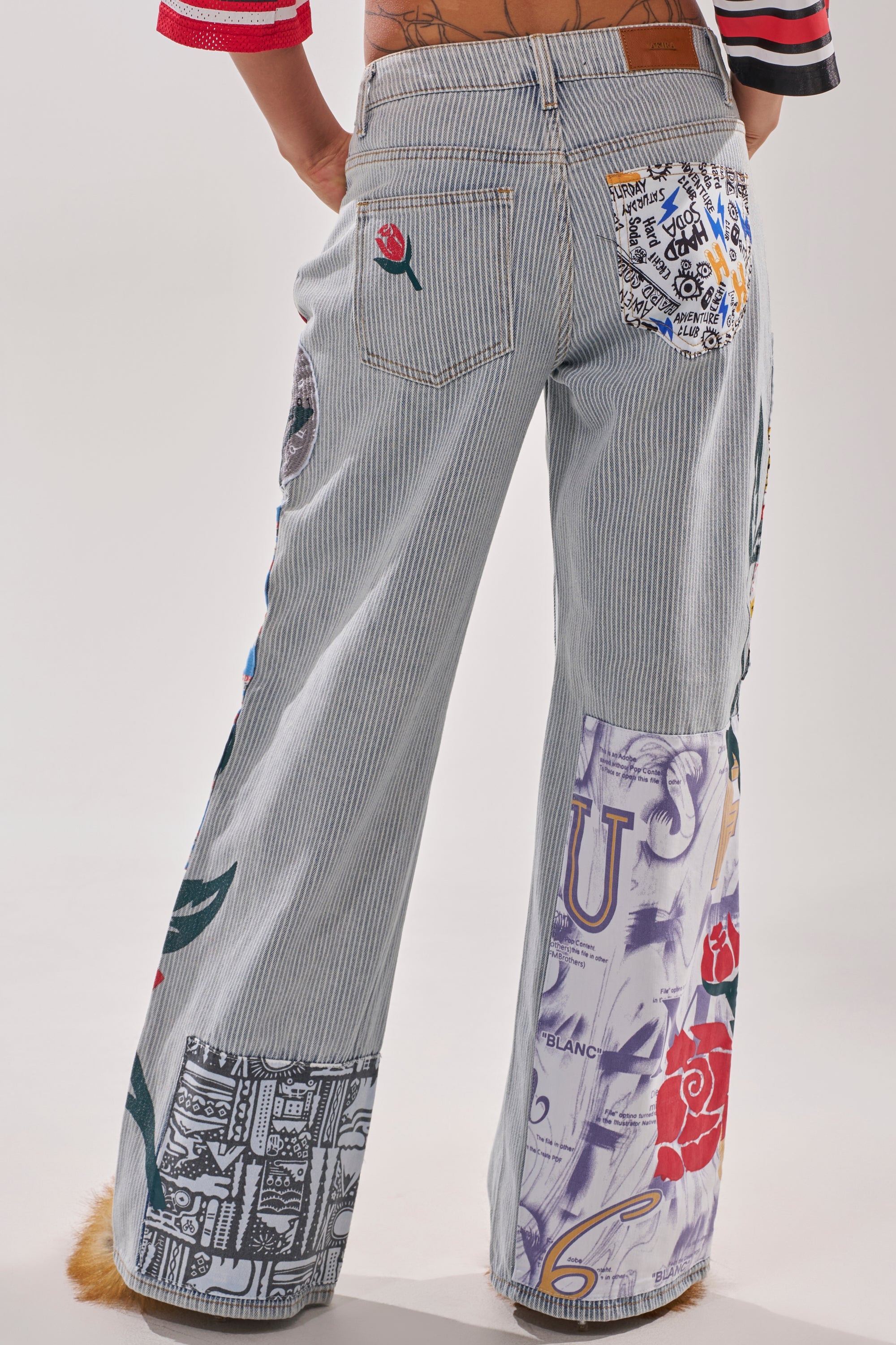 A person stands with their back to the camera wearing the CIAO BELLA STRIPE TROUSER—high-rise, wide-leg jeans in gray and white pinstripes, adorned with colorful patchwork and graphic prints. 4