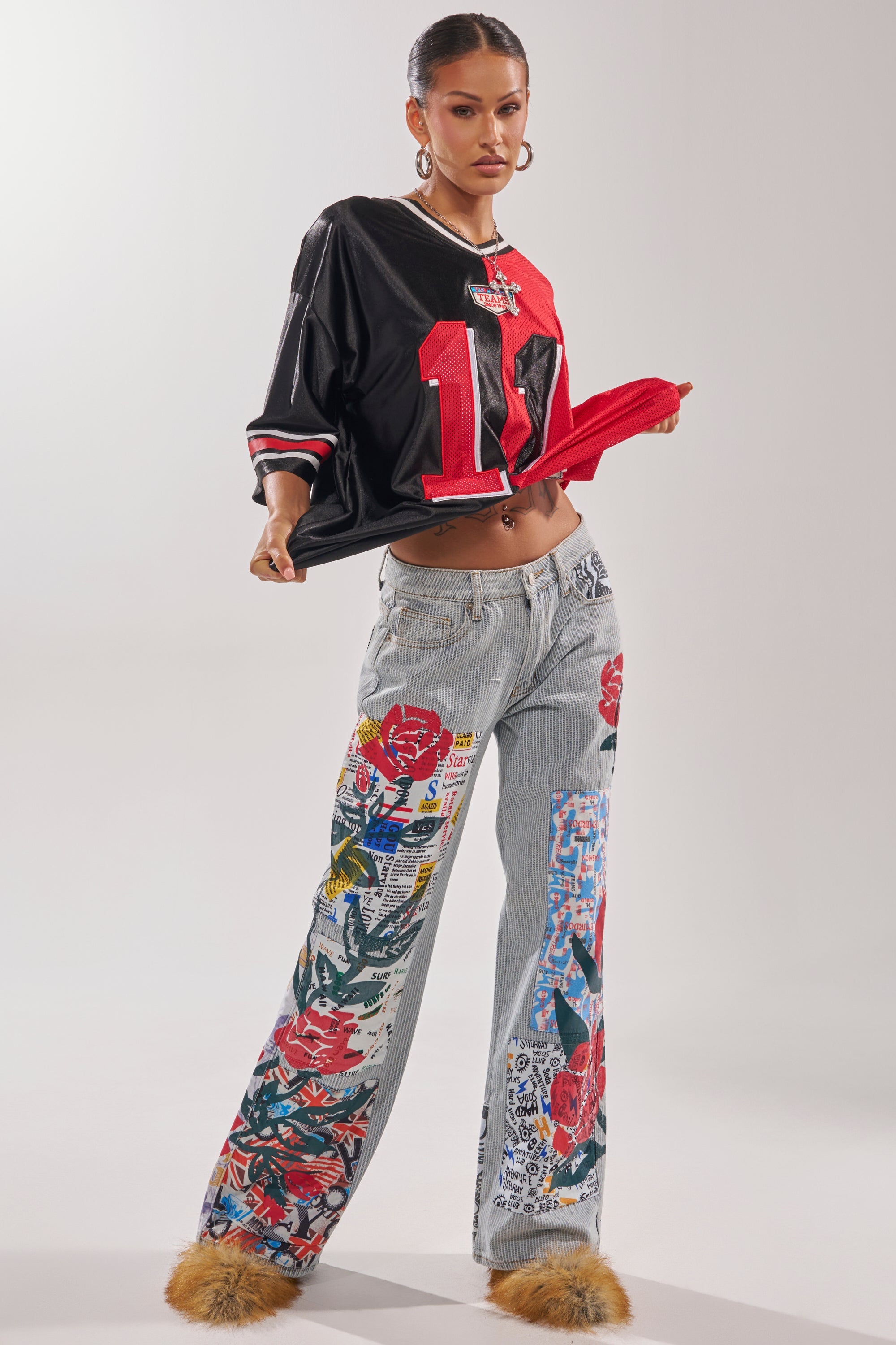 A woman poses confidently against a plain background, wearing the CIAO BELLA STRIPE TROUSER, a loose black and red sports jersey with the number 14, large hoop earrings, and fluffy brown slippers. 7