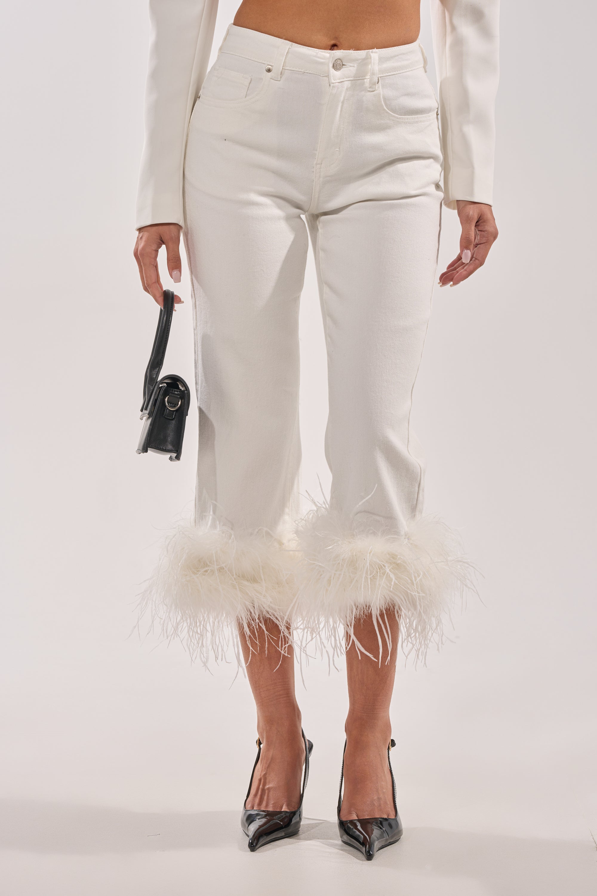A person wearing the TRIPPY LIKE ME DENIM PANT WITH FEATHER, a white jacket, black pointed-toe heels, and holding a small black handbag stands against a plain white background. 0