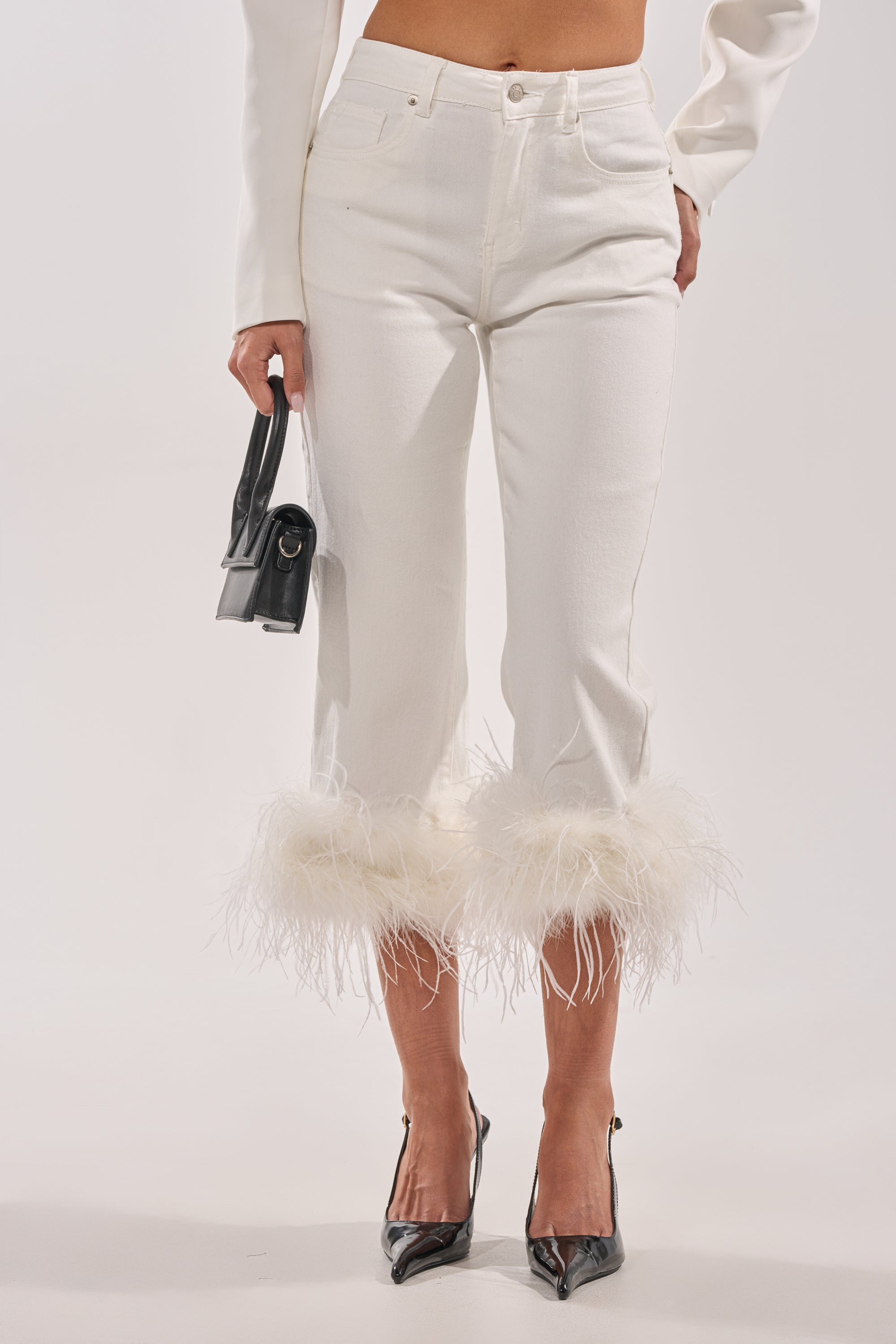 A person models the TRIPPY LIKE ME DENIM PANT WITH FEATHER—high-rise white jeans with feathered hems—styled with a white long-sleeve top, black pointed-toe heels, and a small black handbag against a plain white background. 1