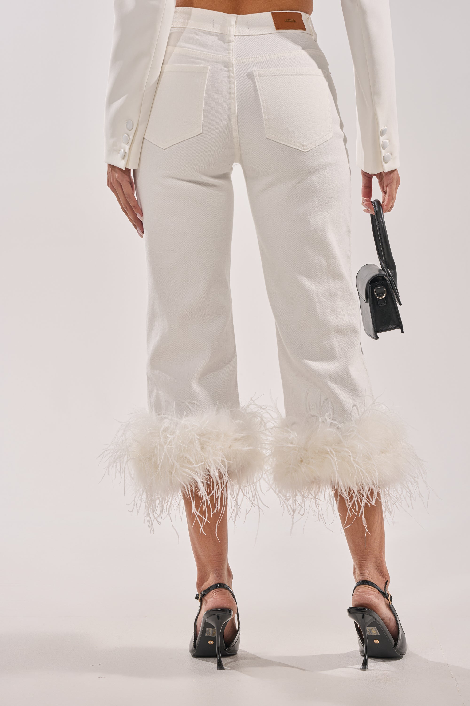 A person is shown from the back wearing TRIPPY LIKE ME DENIM PANT WITH FEATHER (white, high-rise), a white blazer, black heeled sandals, and holding a small black handbag against a light background. 3