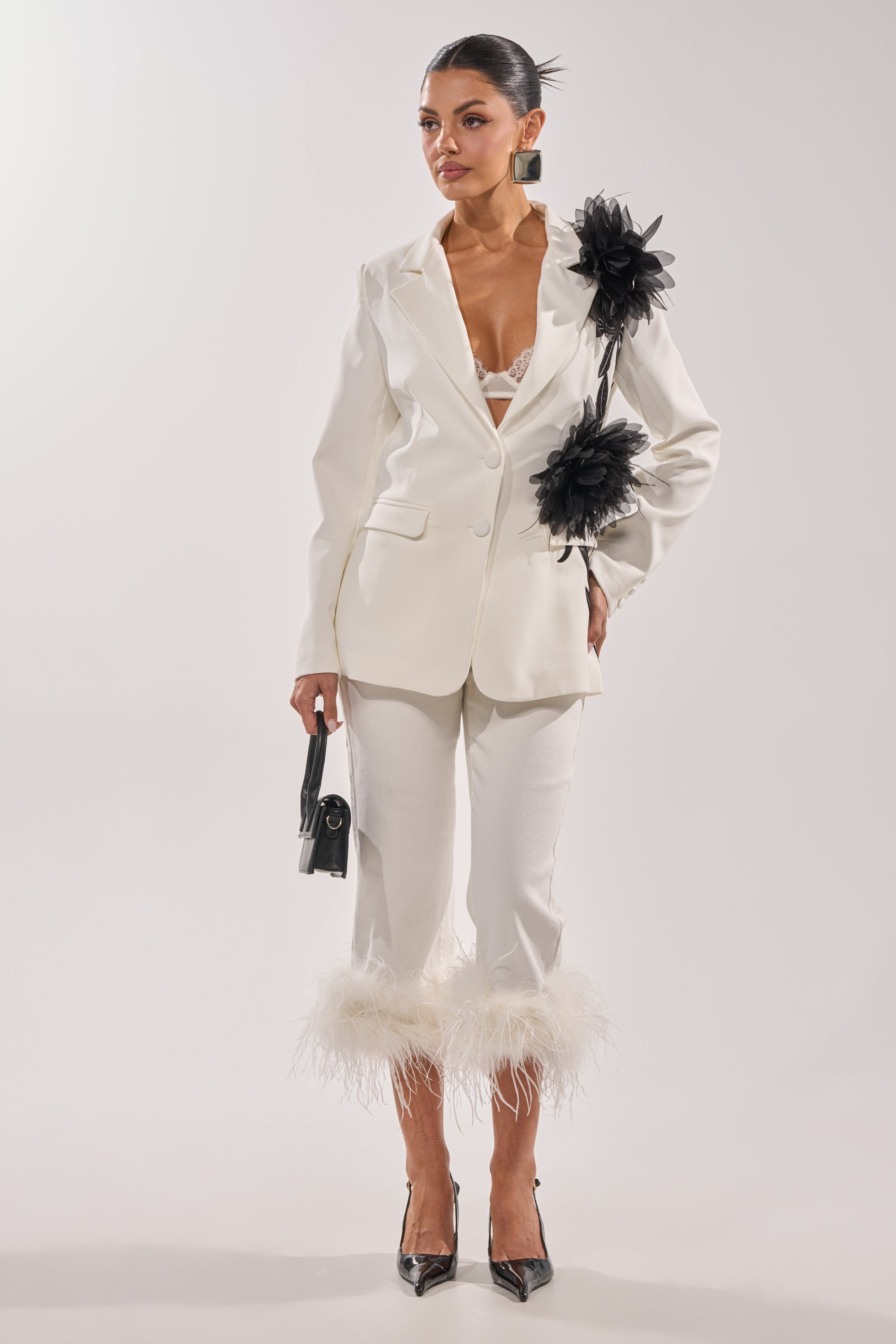 A woman stands against a plain background wearing the TRIPPY LIKE ME DENIM PANT WITH FEATHER in white, paired with a white blazer featuring black floral accents, black high heels, and holding a small black handbag. 5