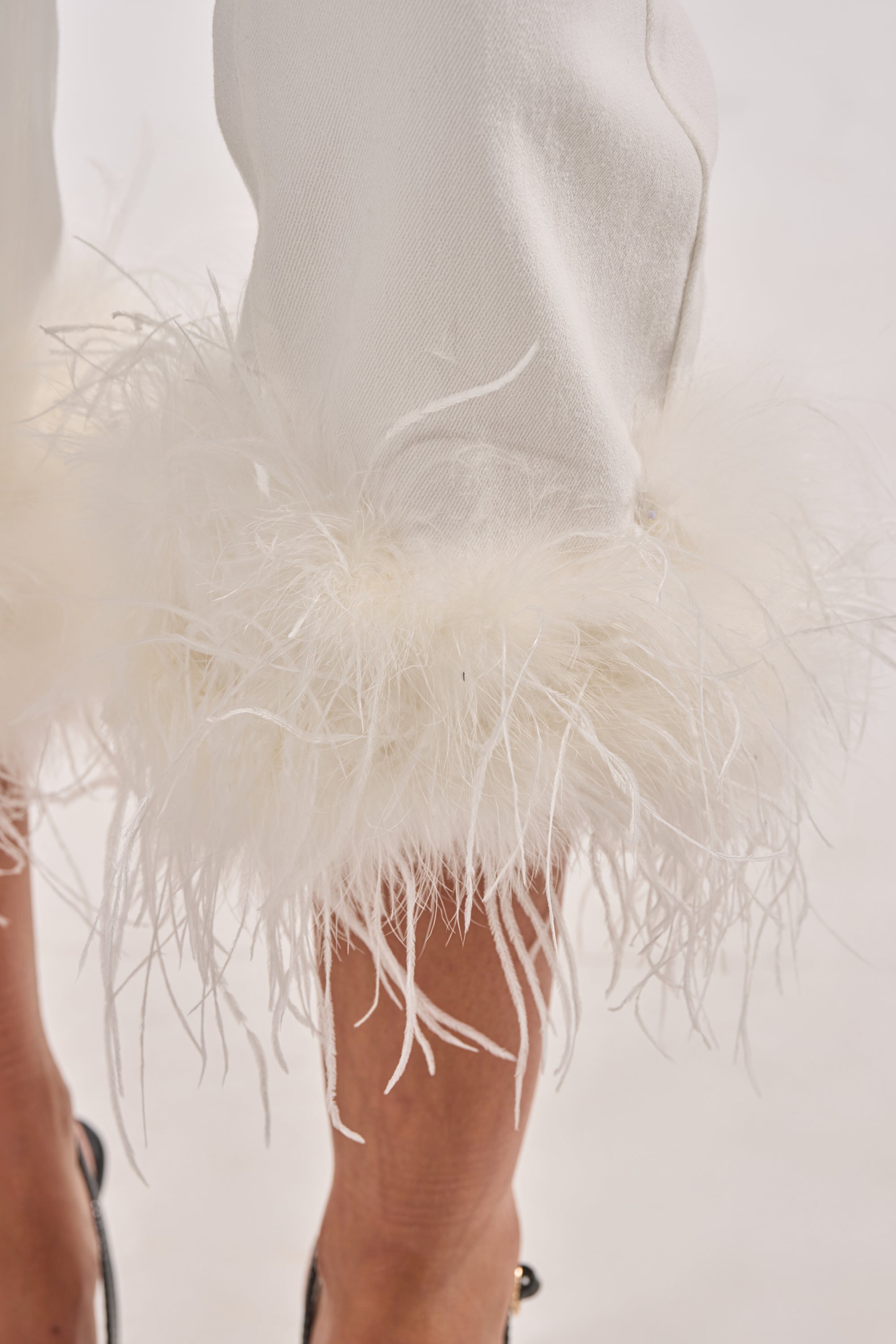 Close-up of the TRIPPY LIKE ME DENIM PANT WITH FEATHER, featuring white high-rise straight-leg jeans with fluffy feather-trimmed cuffs, styled with black sandals against a light neutral background. 6