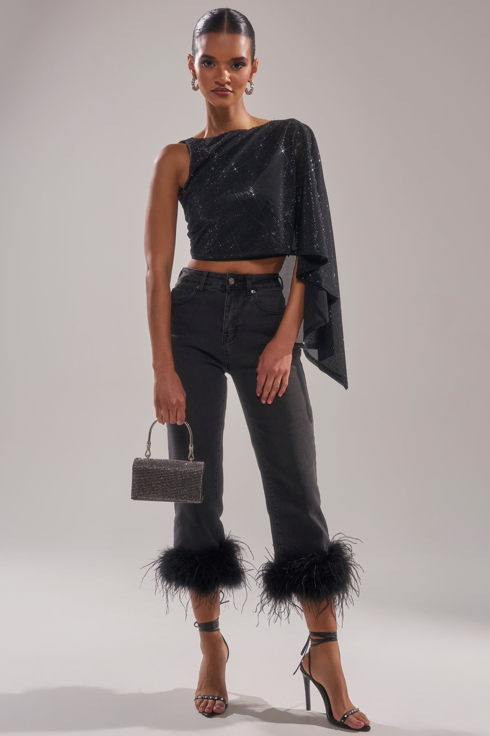 A woman models the TRIPPY LIKE ME DENIM PANT WITH FEATHER, paired with a black one-shoulder sparkly top, strappy heeled sandals, glittery handbag, and statement earrings. Her hair is styled back for a polished look. 0