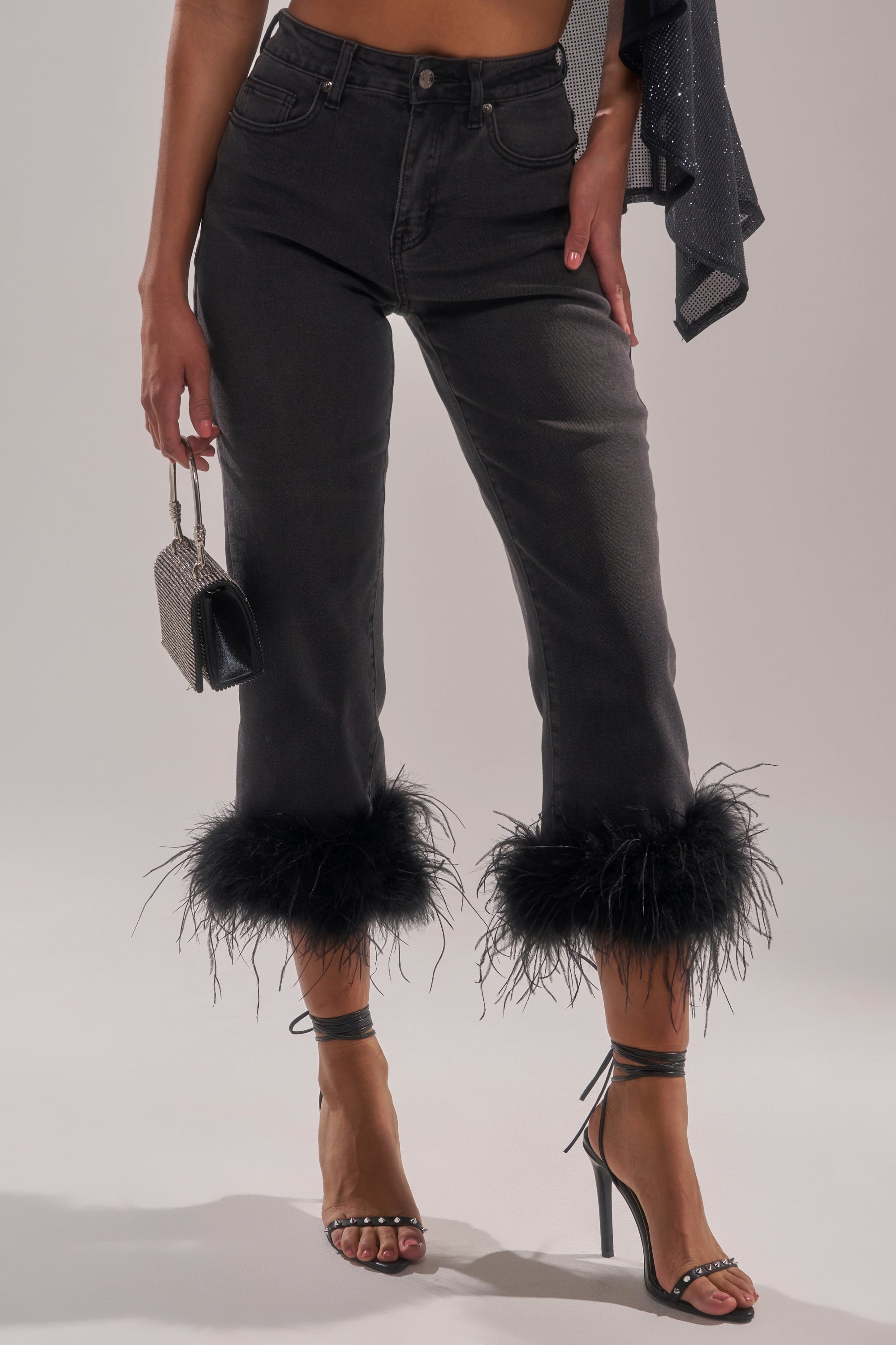 A person wears the TRIPPY LIKE ME DENIM PANT WITH FEATHER, paired with strappy high heels and a small purse. Only their lower body is visible against a neutral background. 1