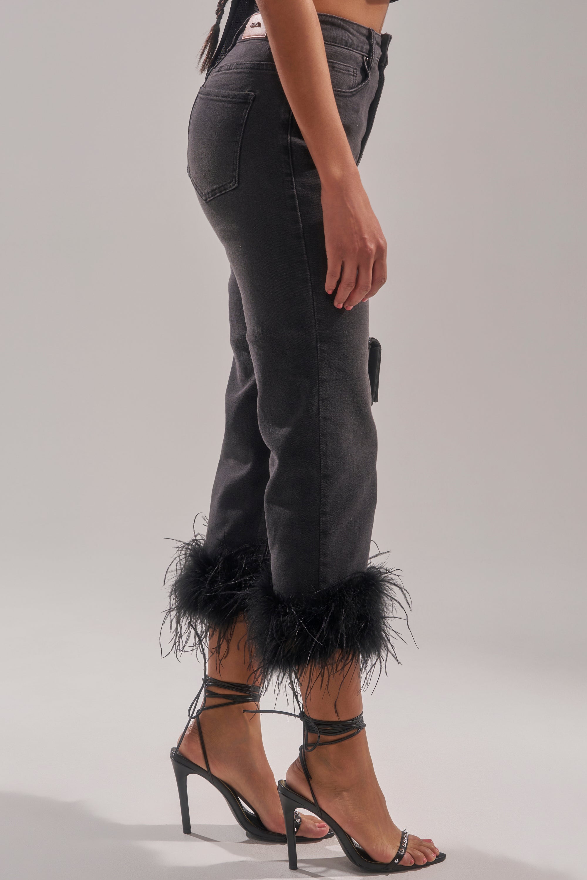 A person wearing the TRIPPY LIKE ME DENIM PANT WITH FEATHER and black strappy high-heeled sandals, photographed from the side against a plain background. 3