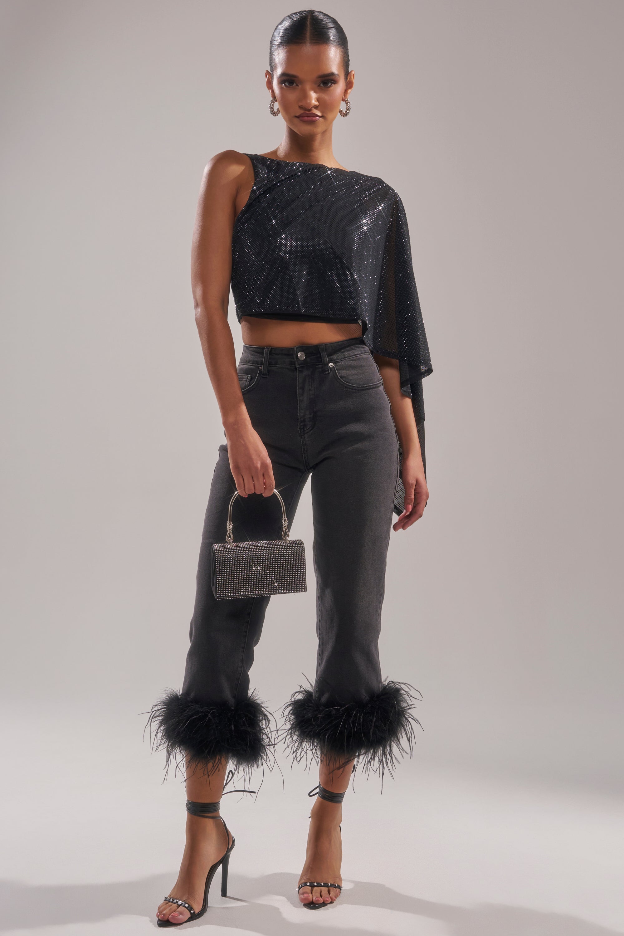 A woman wears a black, one-shoulder sparkly crop top, TRIPPY LIKE ME DENIM PANT WITH FEATHER, strappy high heels, and holds a small glittery handbag. 6