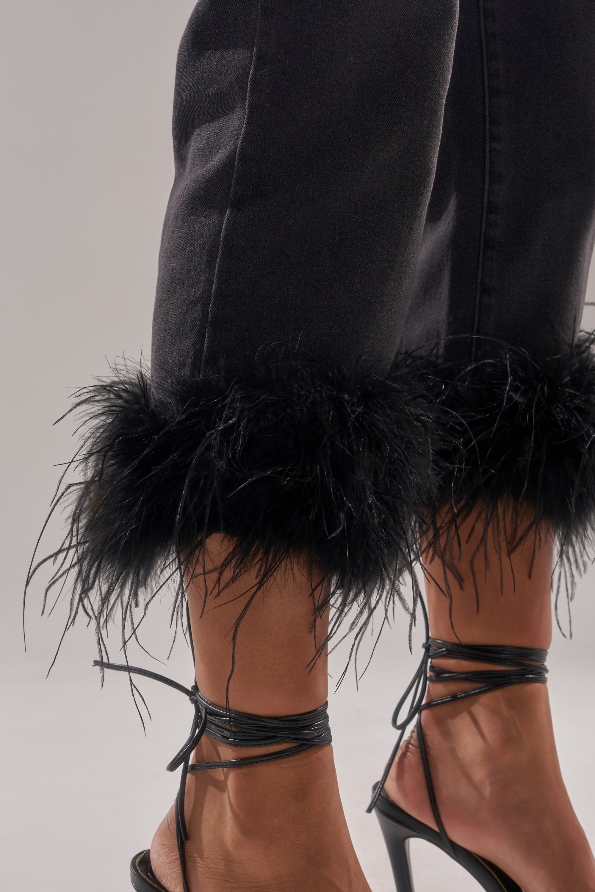 Close-up of a person's lower legs wearing the TRIPPY LIKE ME DENIM PANT WITH FEATHER and black strappy high-heeled sandals against a plain light background. 7