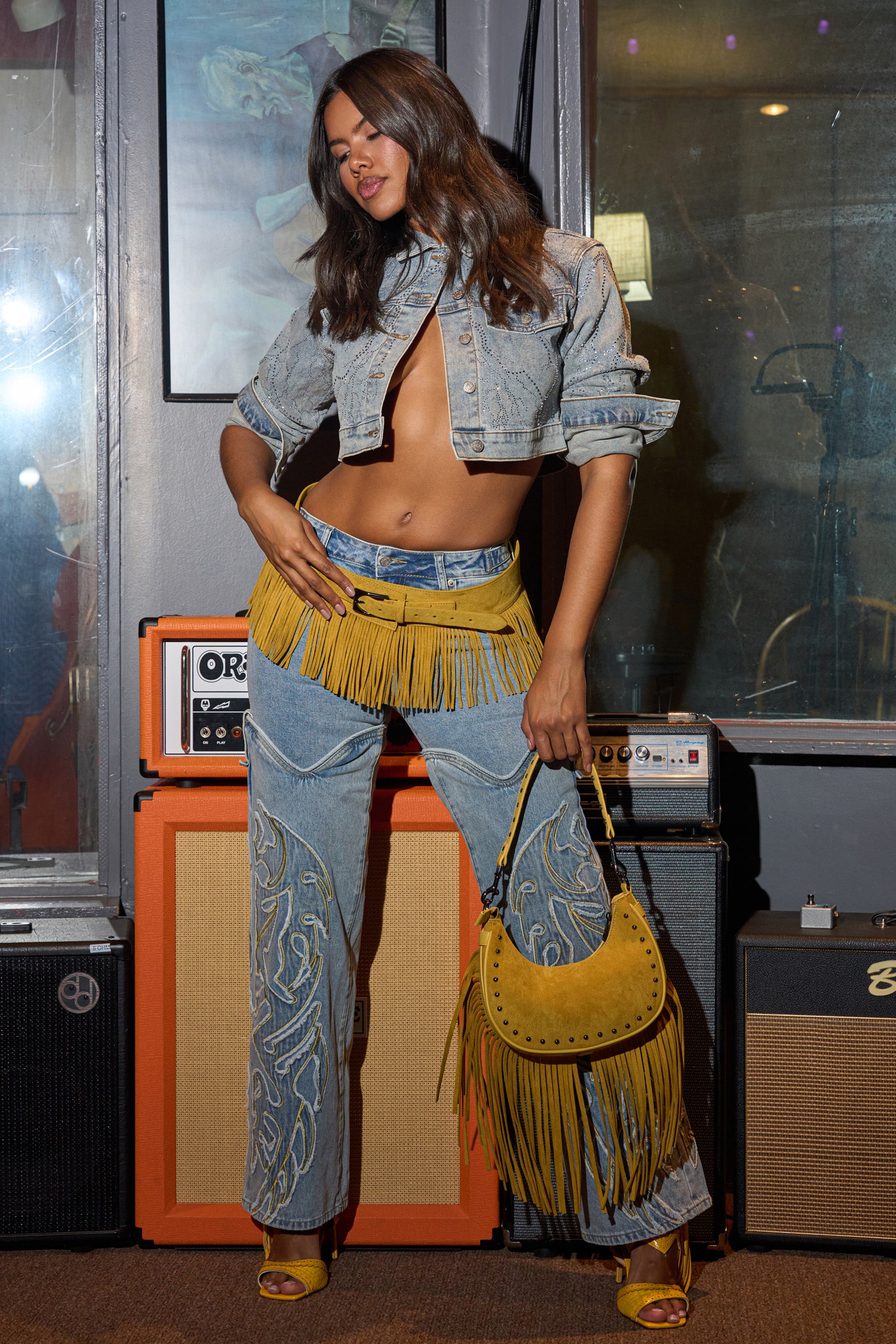 A woman poses indoors in streetwear, wearing BUCKIN' EMBROIDERED DENIM JEANS with yellow fringe, a cropped denim jacket, matching heeled sandals, and a yellow fringed handbag; guitar amps sit behind her.