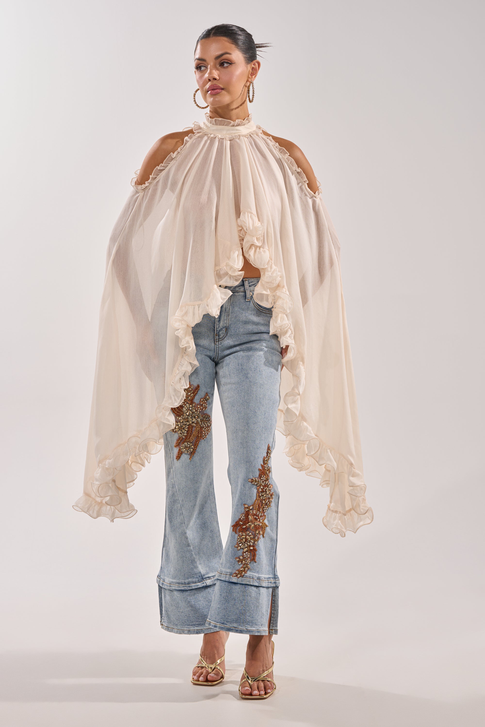 A woman models the TOTALLY COOL RELAXED DENIM jeans with a sheer, off-white ruffled cape top, gold heels, and hoop earrings. Her hair is styled up. 3