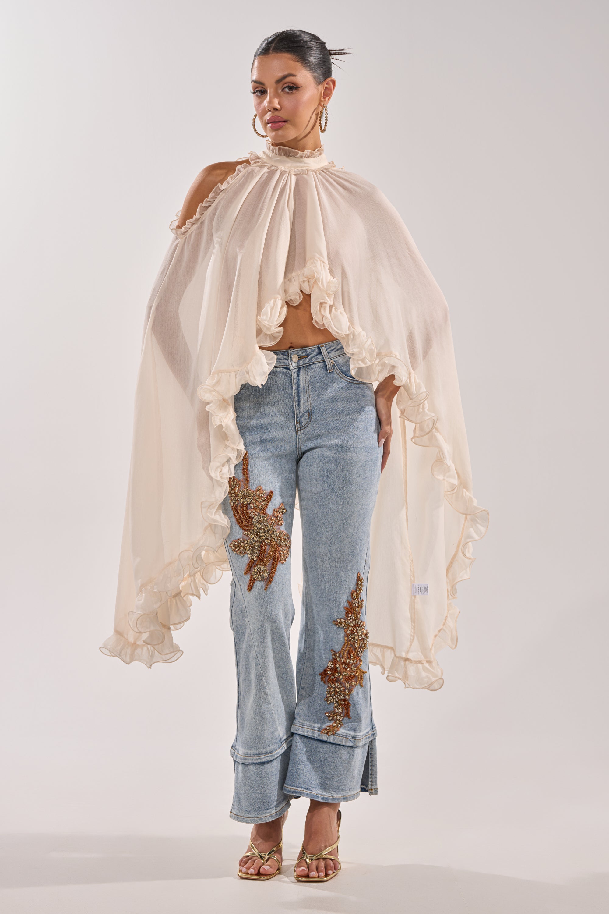 A woman stands wearing a flowing, sheer ivory cape top with ruffled edges, TOTALLY COOL RELAXED DENIM high-rise jeans adorned with brown floral appliqué, gold hoop earrings, and gold open-toe heels. 4