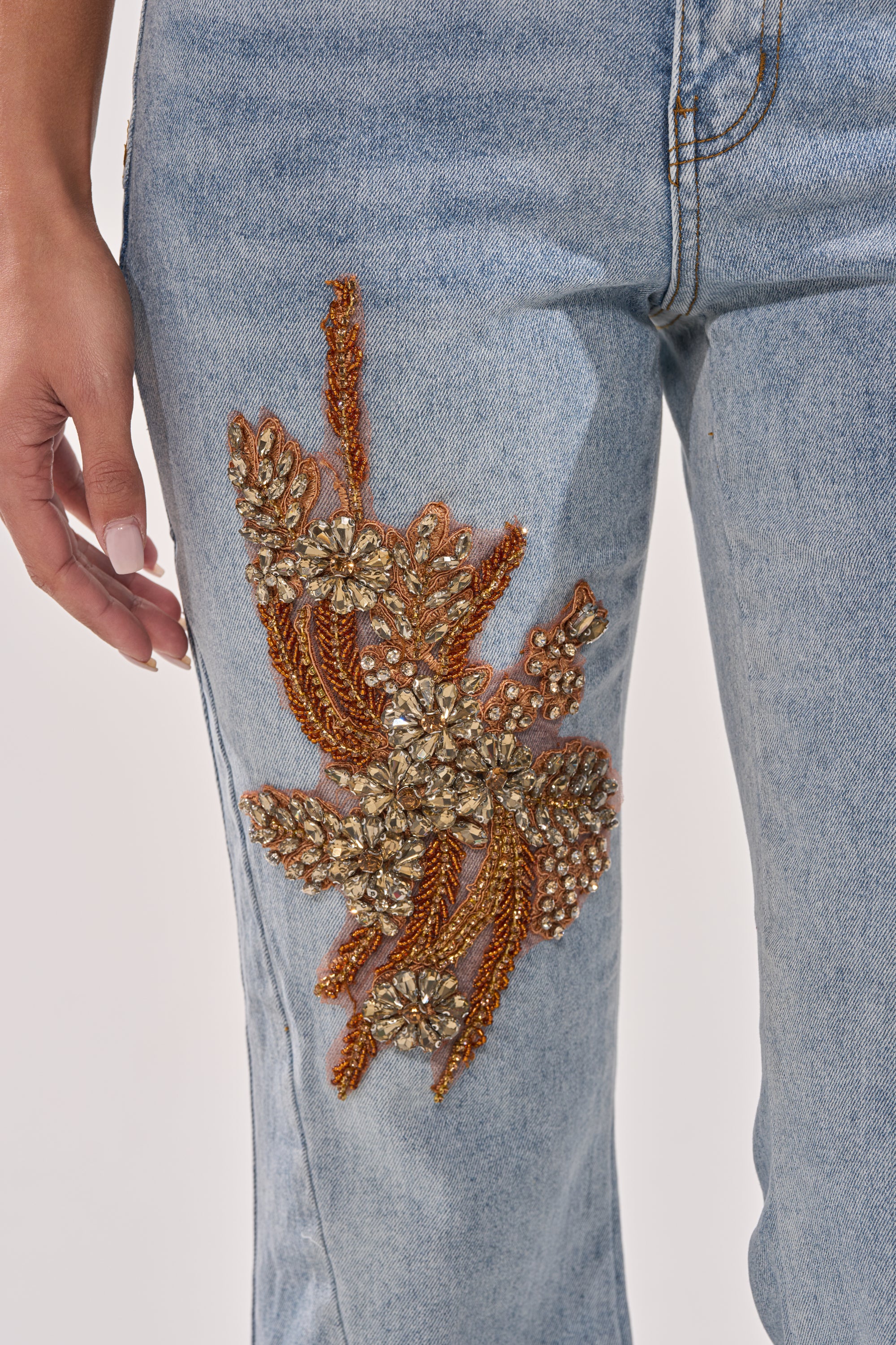 A close-up of TOTALLY COOL RELAXED DENIM: light blue, high-rise jeans adorned with detailed gold and bronze floral embroidery and beadwork on the upper thigh. A manicured hand rests beside the decorated area. 6
