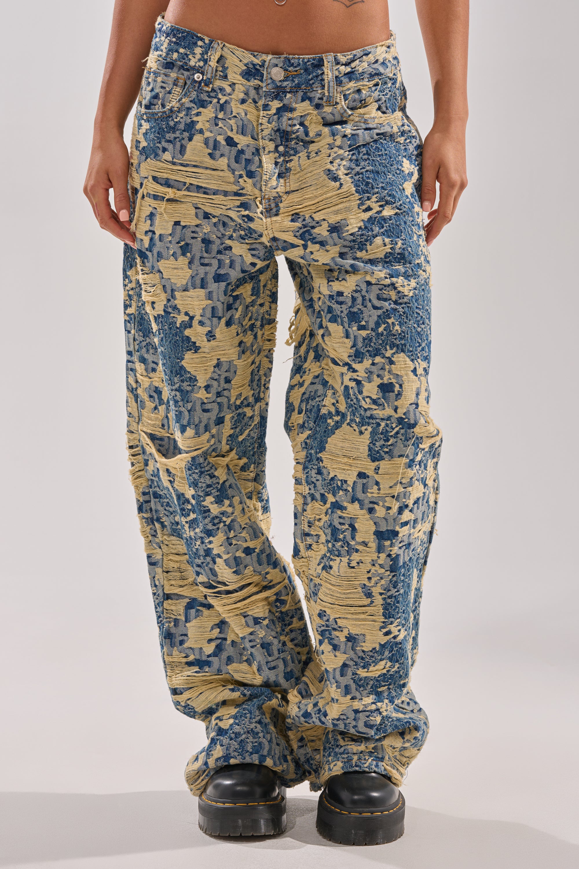 A person models the RENEWED DREAM TAPESTRY PANT—wide-leg, distressed jeans with a blue and yellow abstract pattern—styled with black chunky platform shoes. The image is cropped from mid-torso to feet.