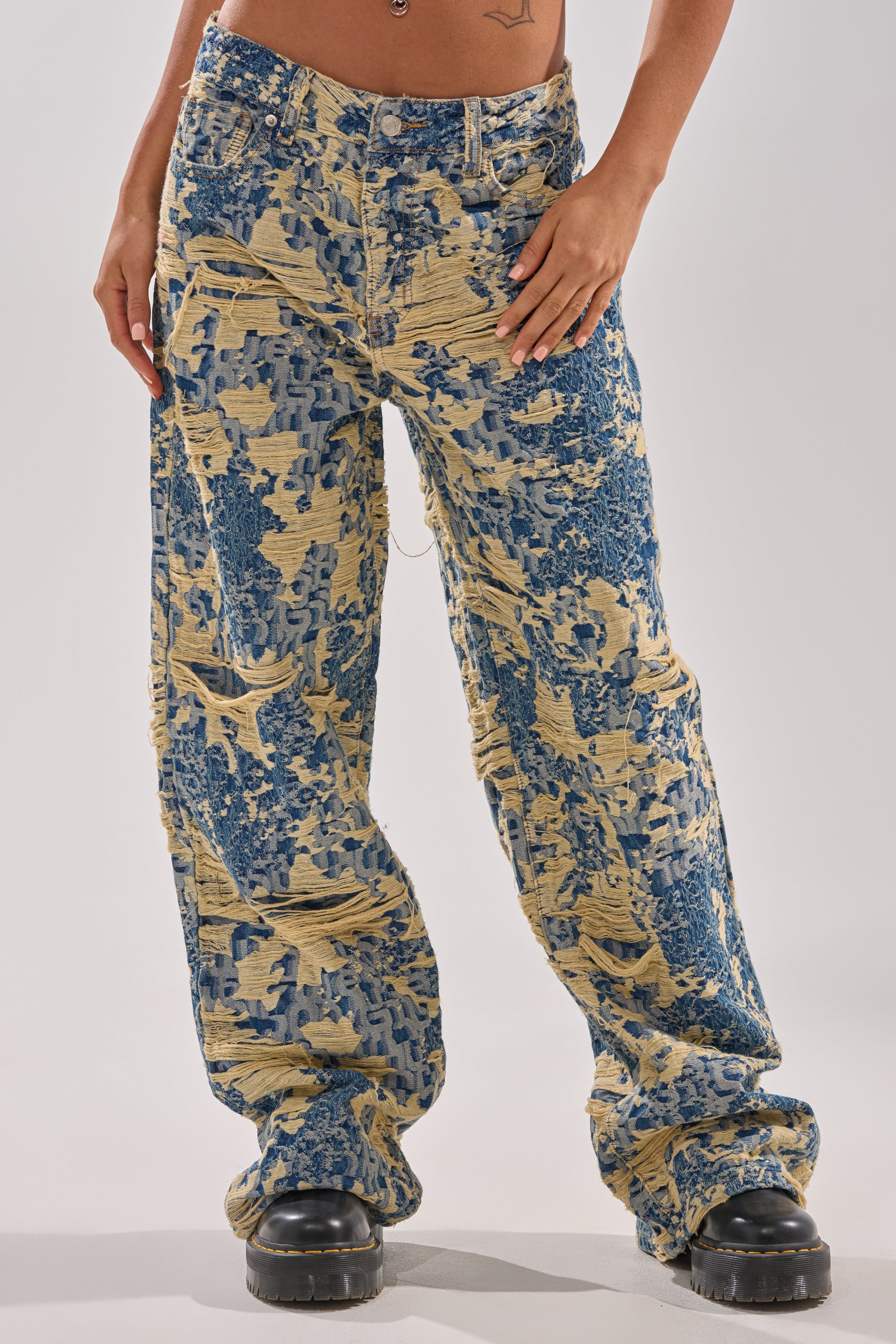 Person wearing RENEWED DREAM TAPESTRY PANT, a high-waisted, wide-leg jean featuring a blue and tan abstract pattern, styled with chunky black platform shoes and a small lower torso tattoo visible.