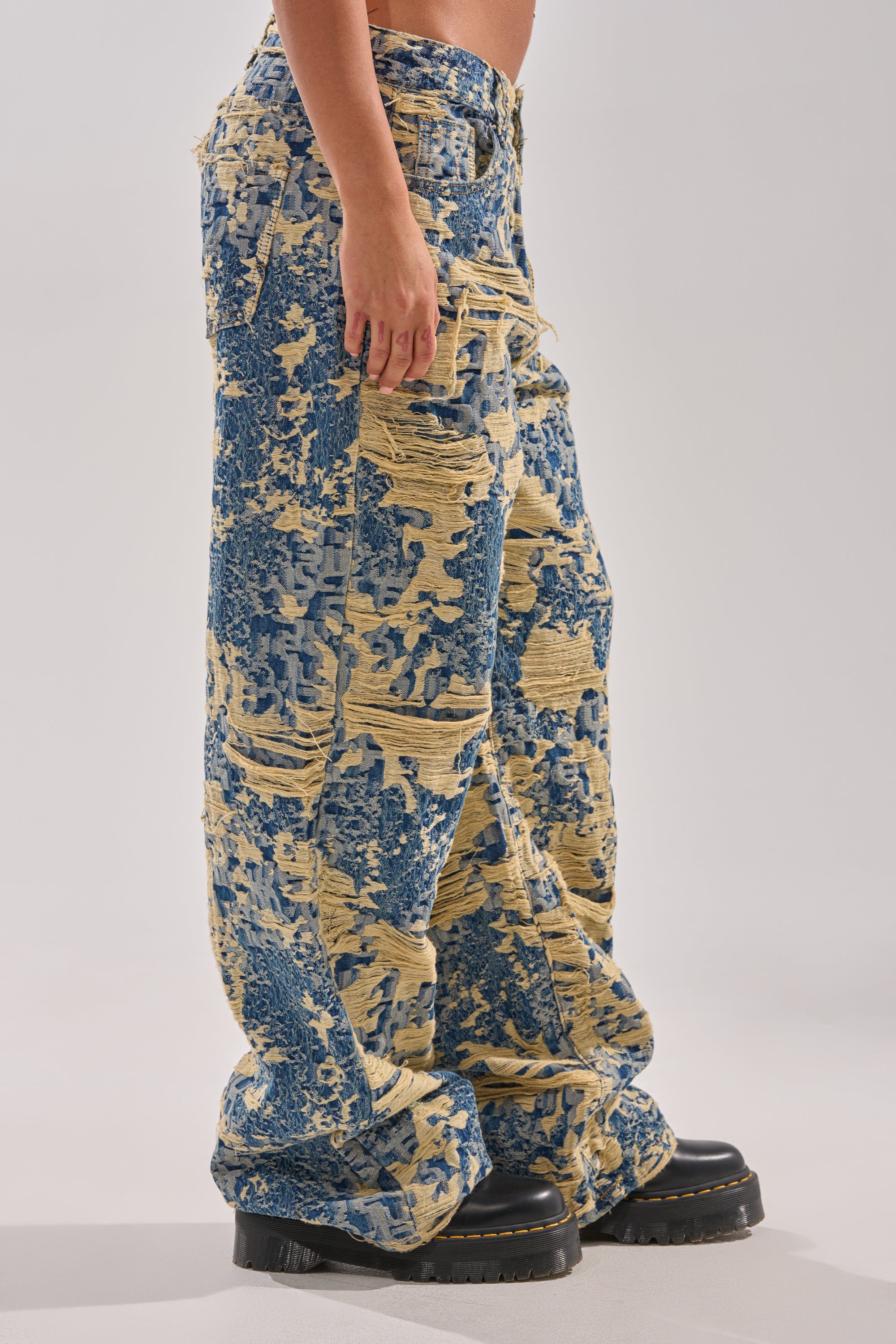 Wearing the RENEWED DREAM TAPESTRY PANT—loose, wide-leg distressed denim in blue and beige patterns with black platform shoes—a person stands against a plain light background, showing only their lower body.