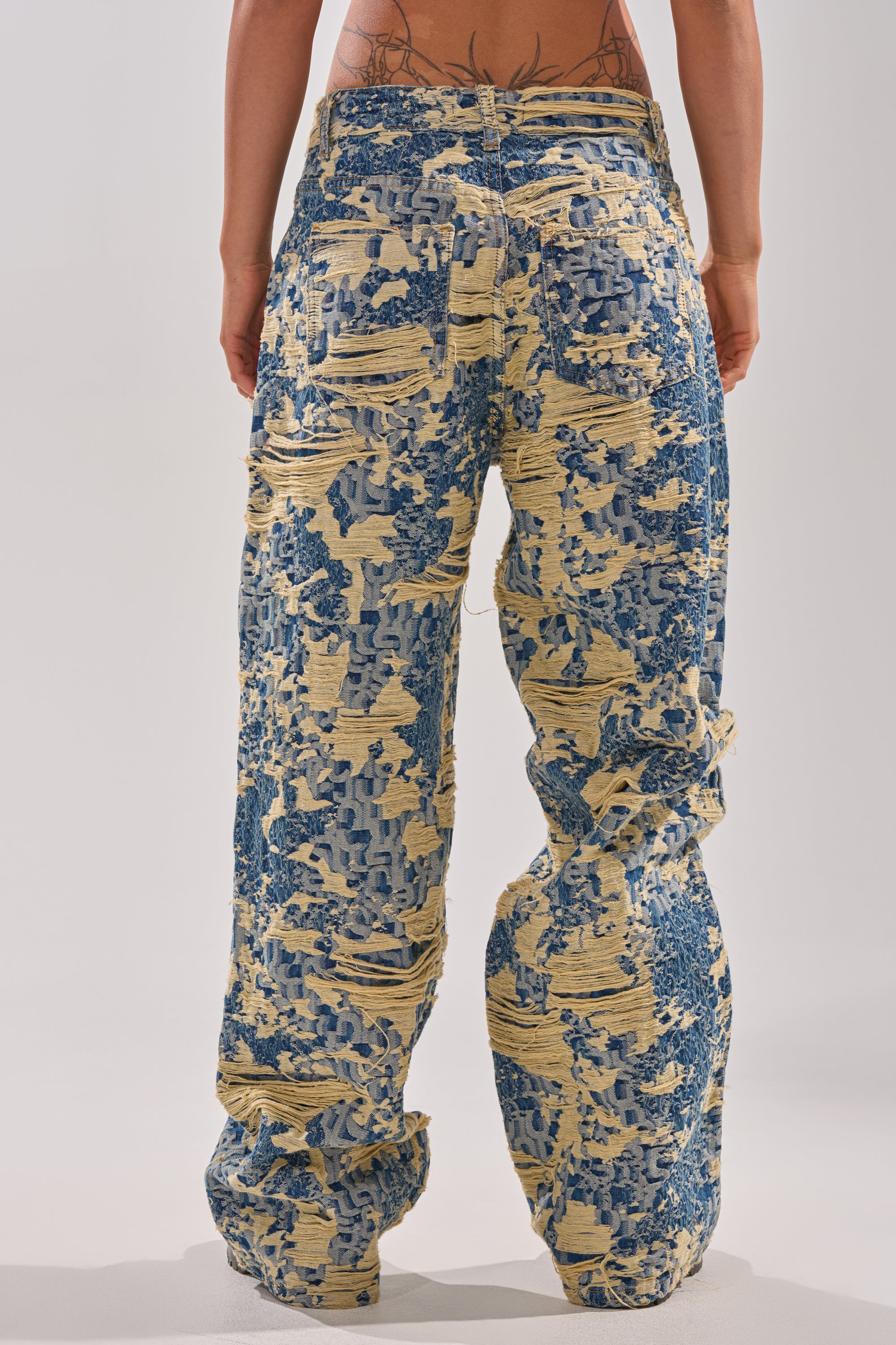 A person stands facing away, showing their lower back above the waistline, wearing the RENEWED DREAM TAPESTRY PANT—loose-fitting, high-waisted denim with a wide-leg silhouette and textured, two-tone pixelated distressing.